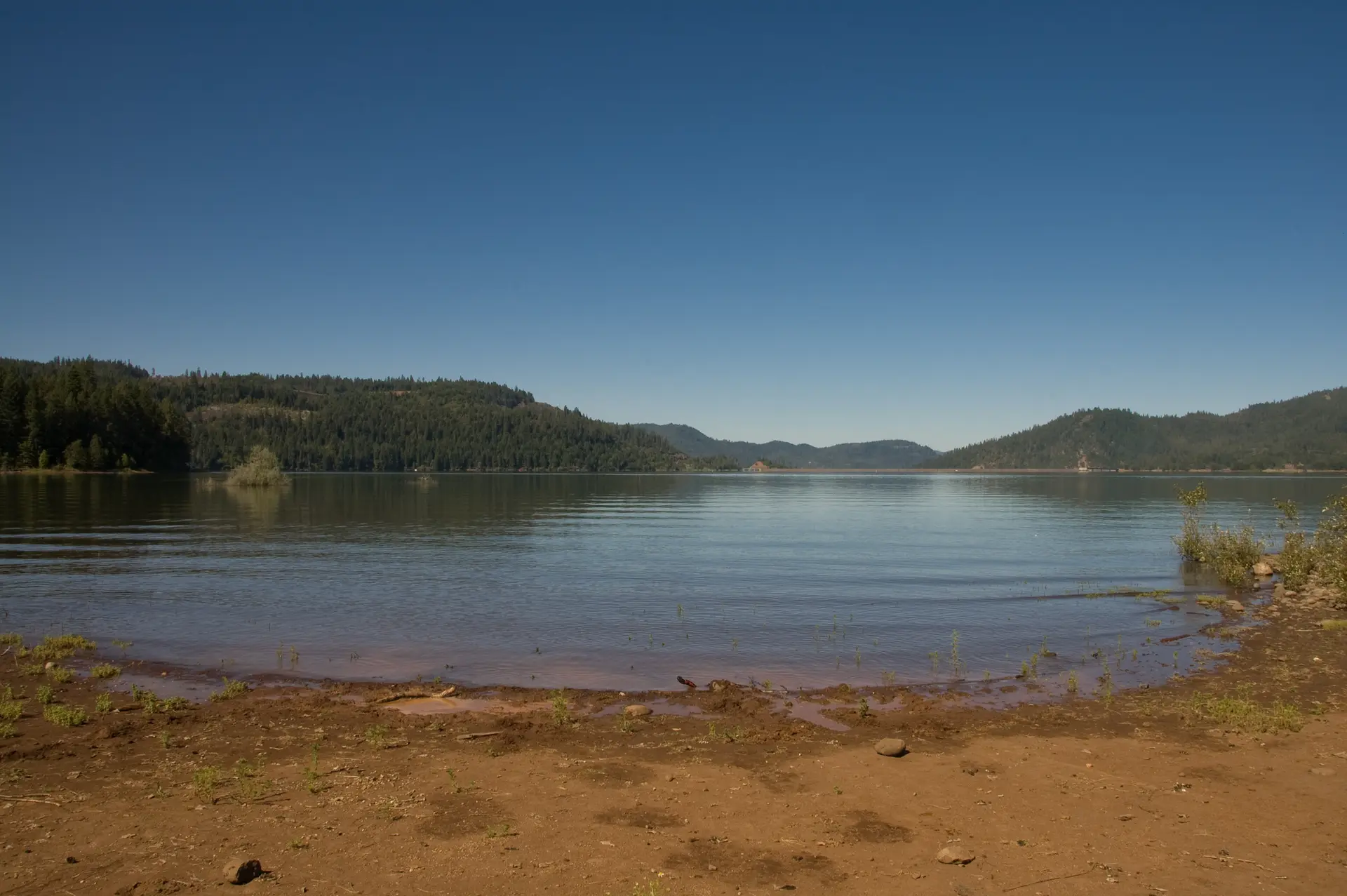 Clear blue sky over a calm lake with forested hills and a sandy shore.