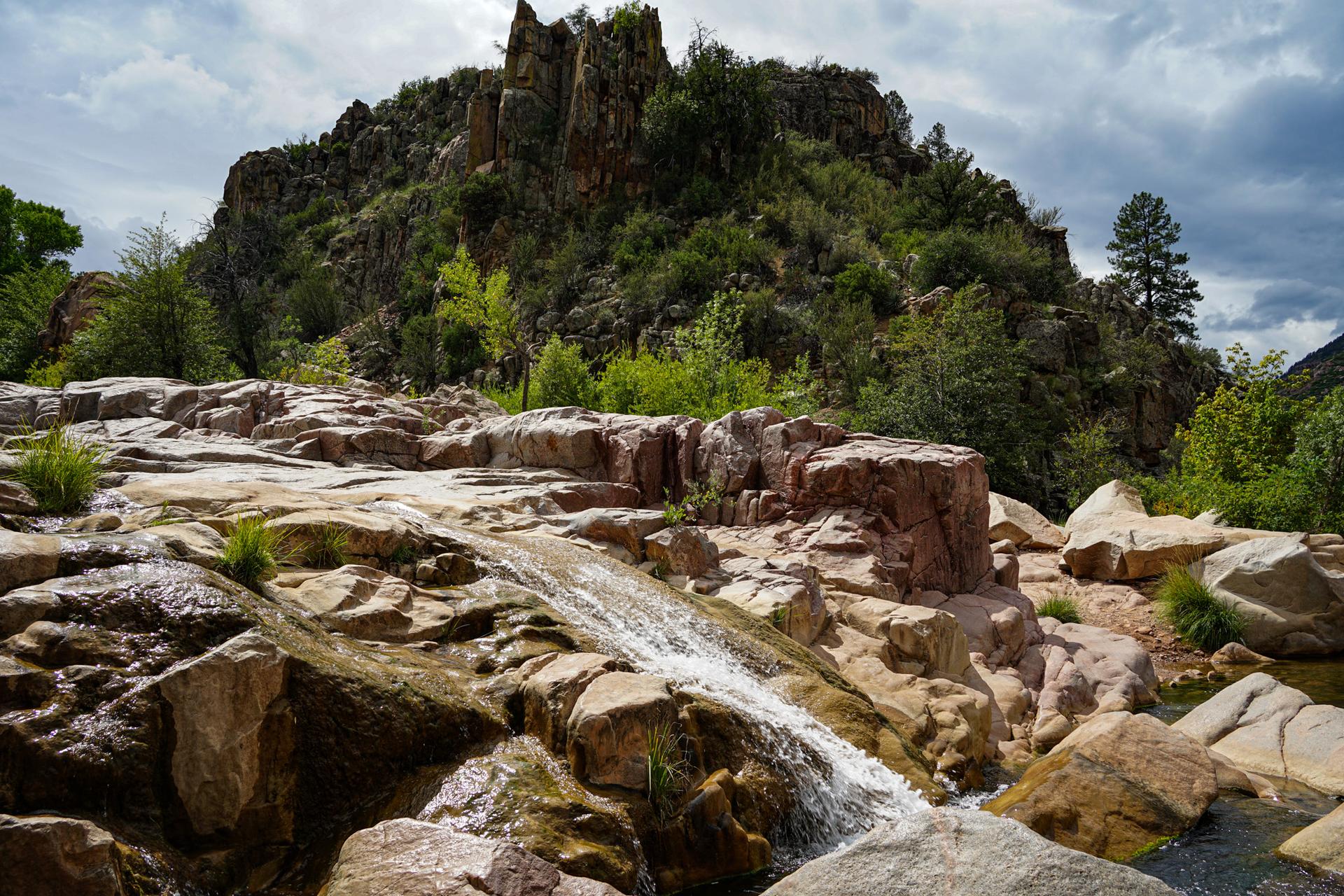 A small waterfall cascades over rocks with a large rocky hill behind.