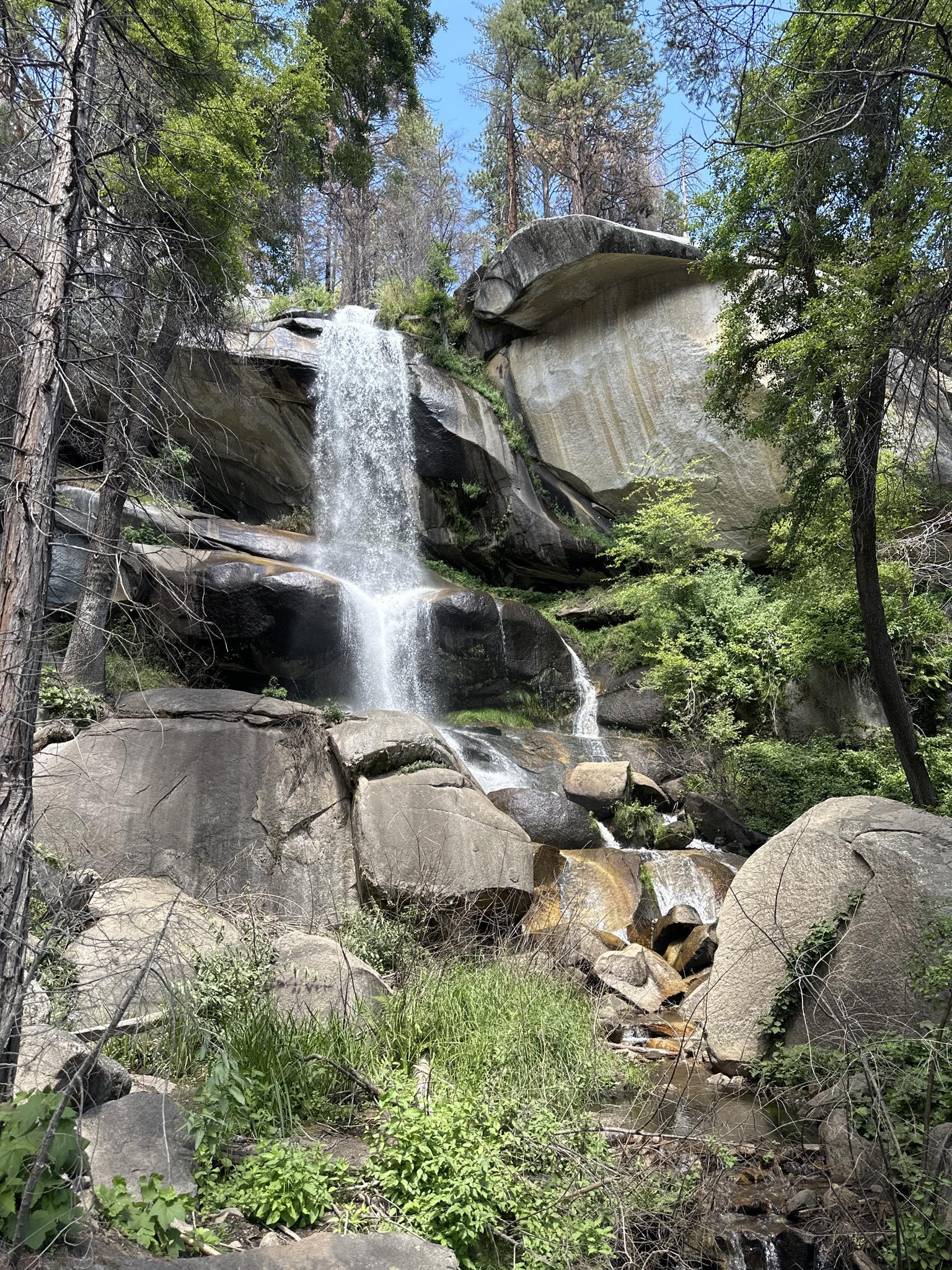 Waterfall cascading over large rocks and green foliage in bright daylight.
