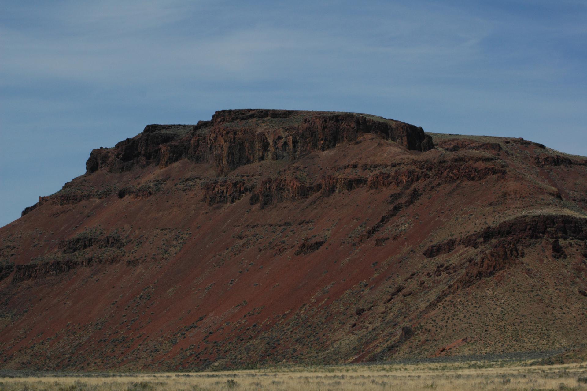 A large, layered, reddish-brown mesa under a bright, partly cloudy sky.