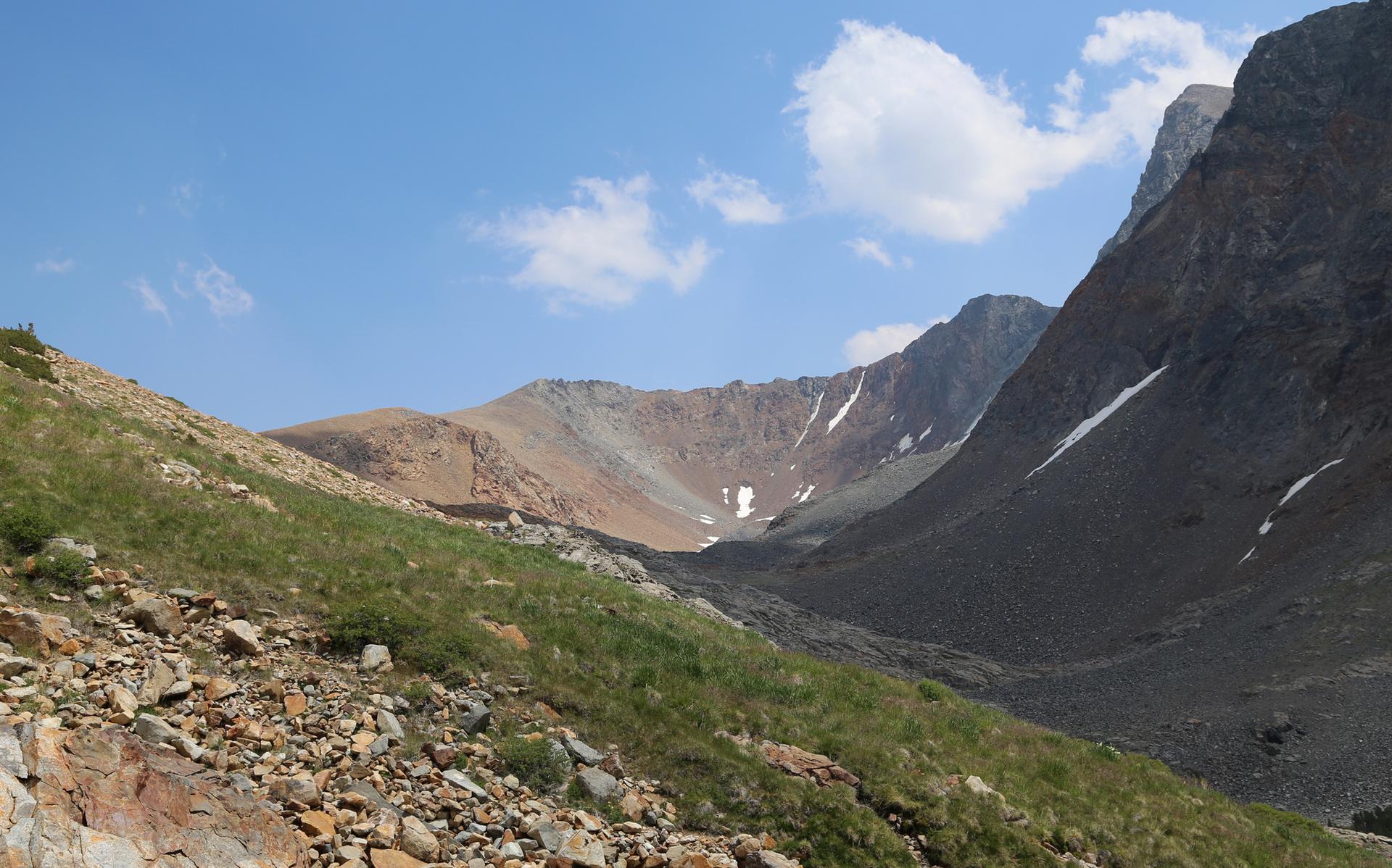 Sunny mountain valley with rocky, grassy slopes and distant peaks under a blue sky.