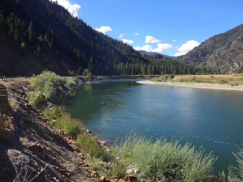 A wide river flows through a sunny mountain valley with forested slopes.