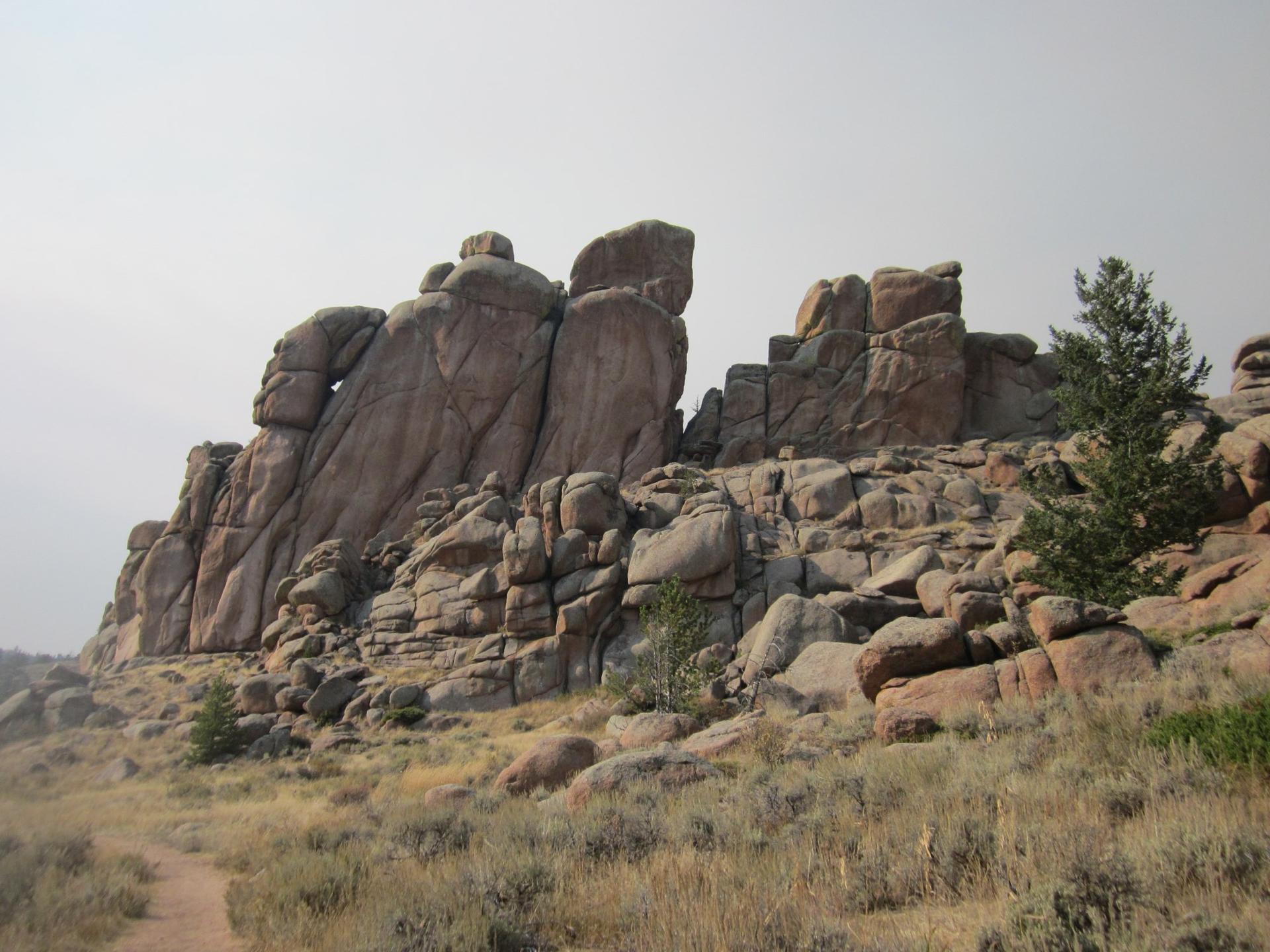 Rocky landscape with massive boulders, sparse trees, and a dirt trail under a hazy sky.