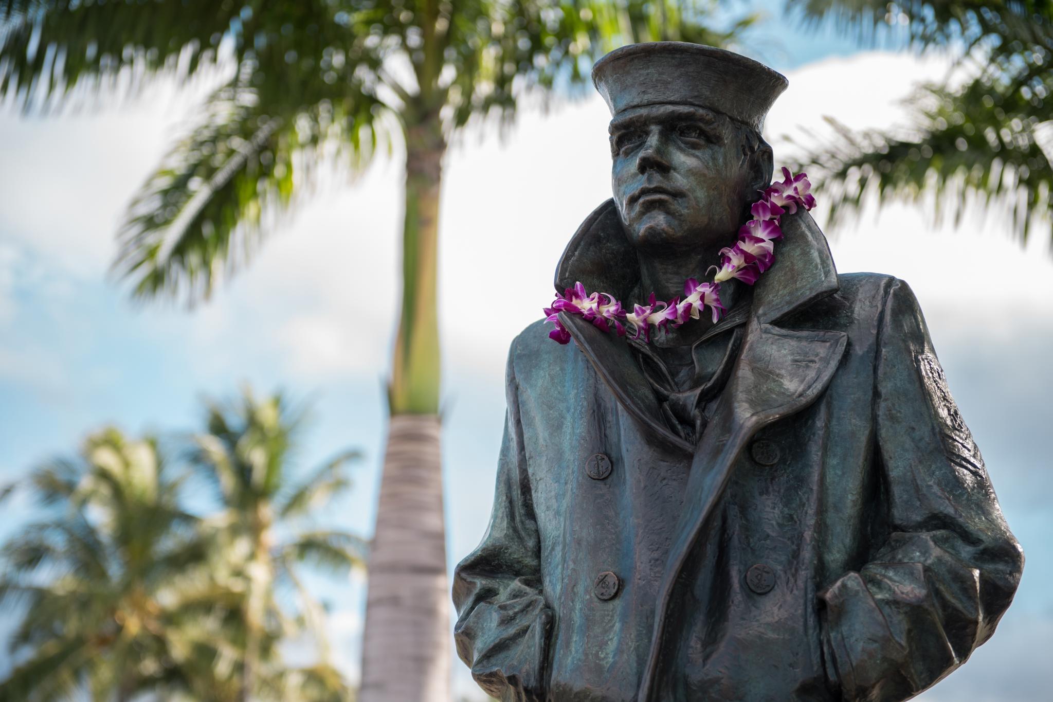 Bronze statue of a sailor wearing a flower lei, with palm trees behind.