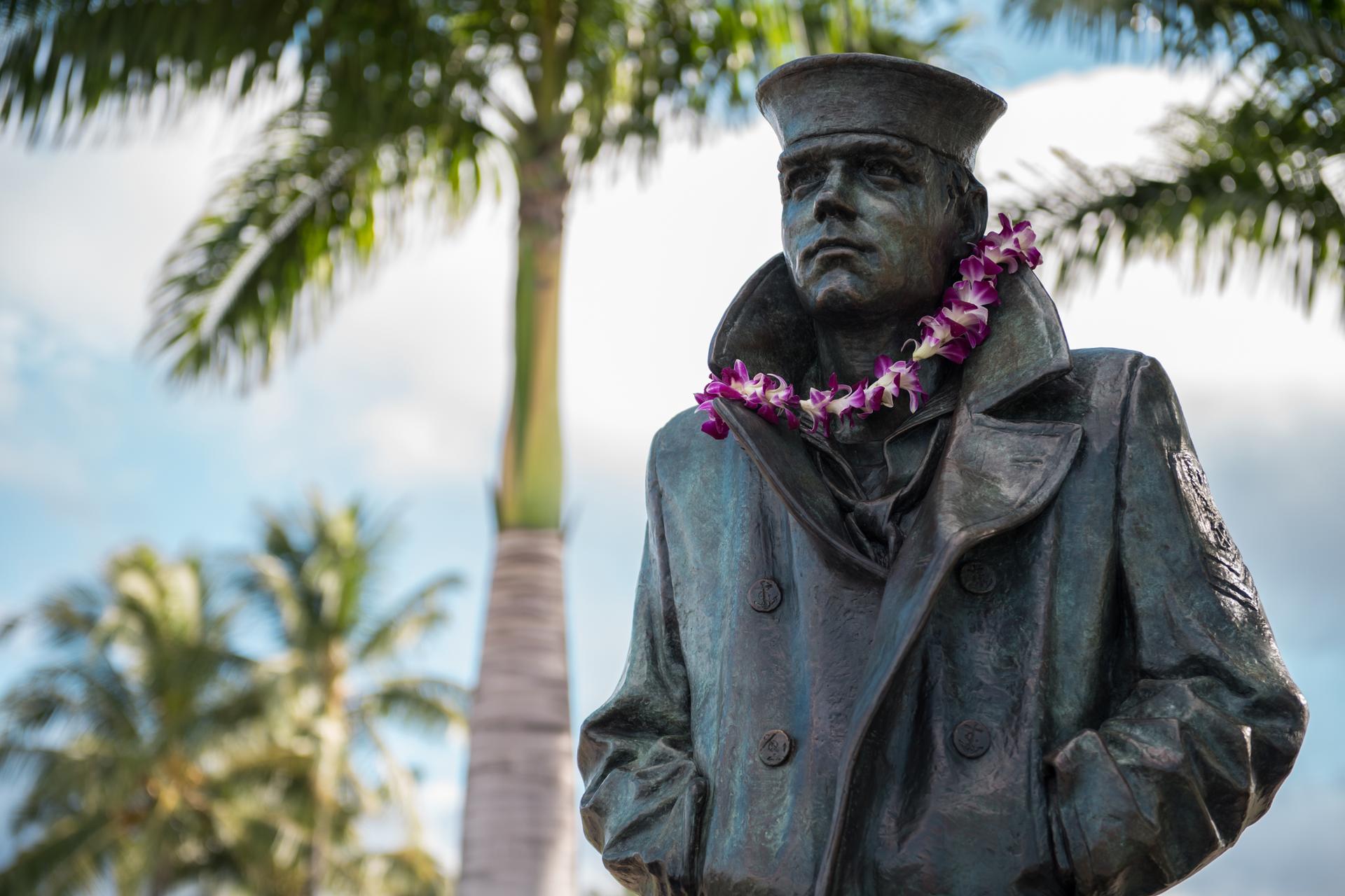 Bronze statue of a sailor wearing a flower lei, with palm trees behind.