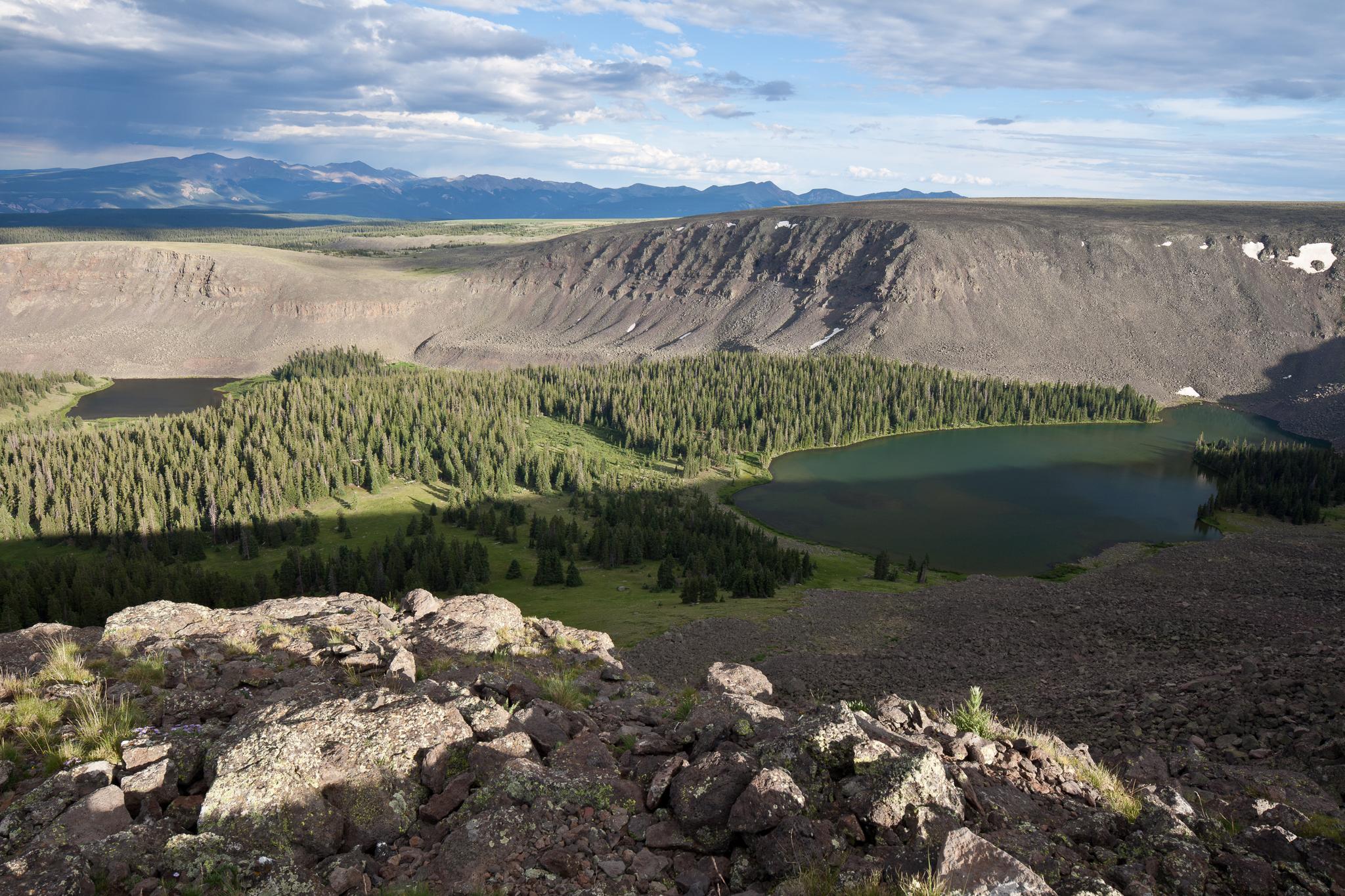 Vast mountain landscape with a lake, evergreen forest, and rocky terrain under clouds.