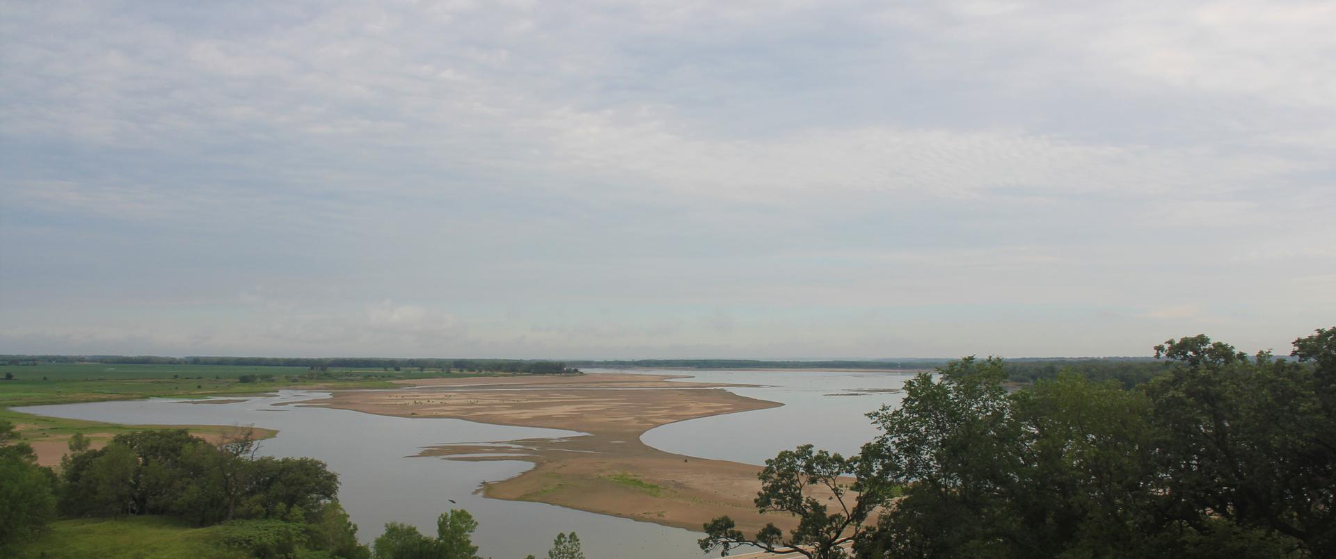 Panoramic view of a wide lake with sandy shores under an overcast sky.