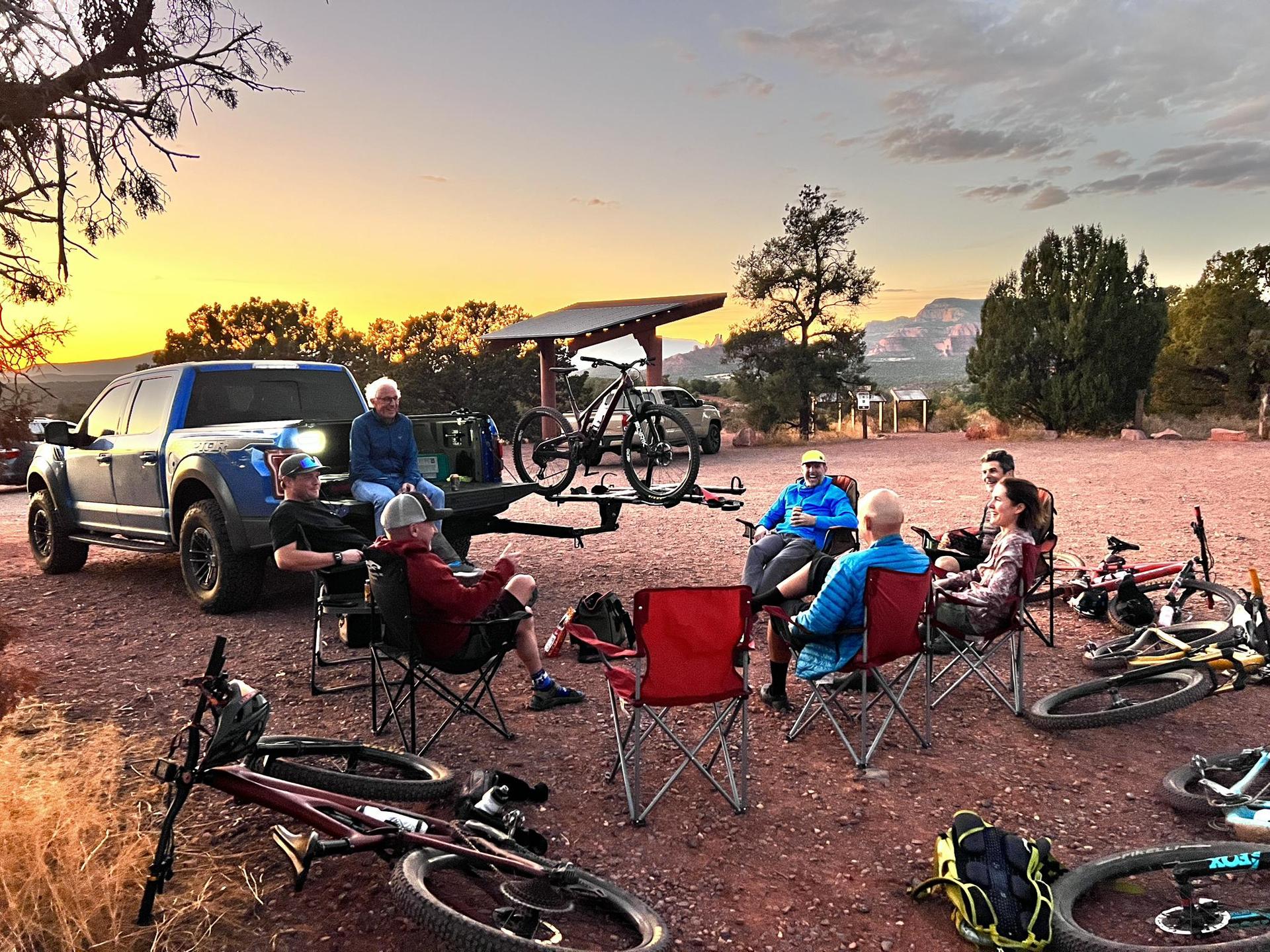 Group of people relaxing in chairs near a truck and bikes at sunset.