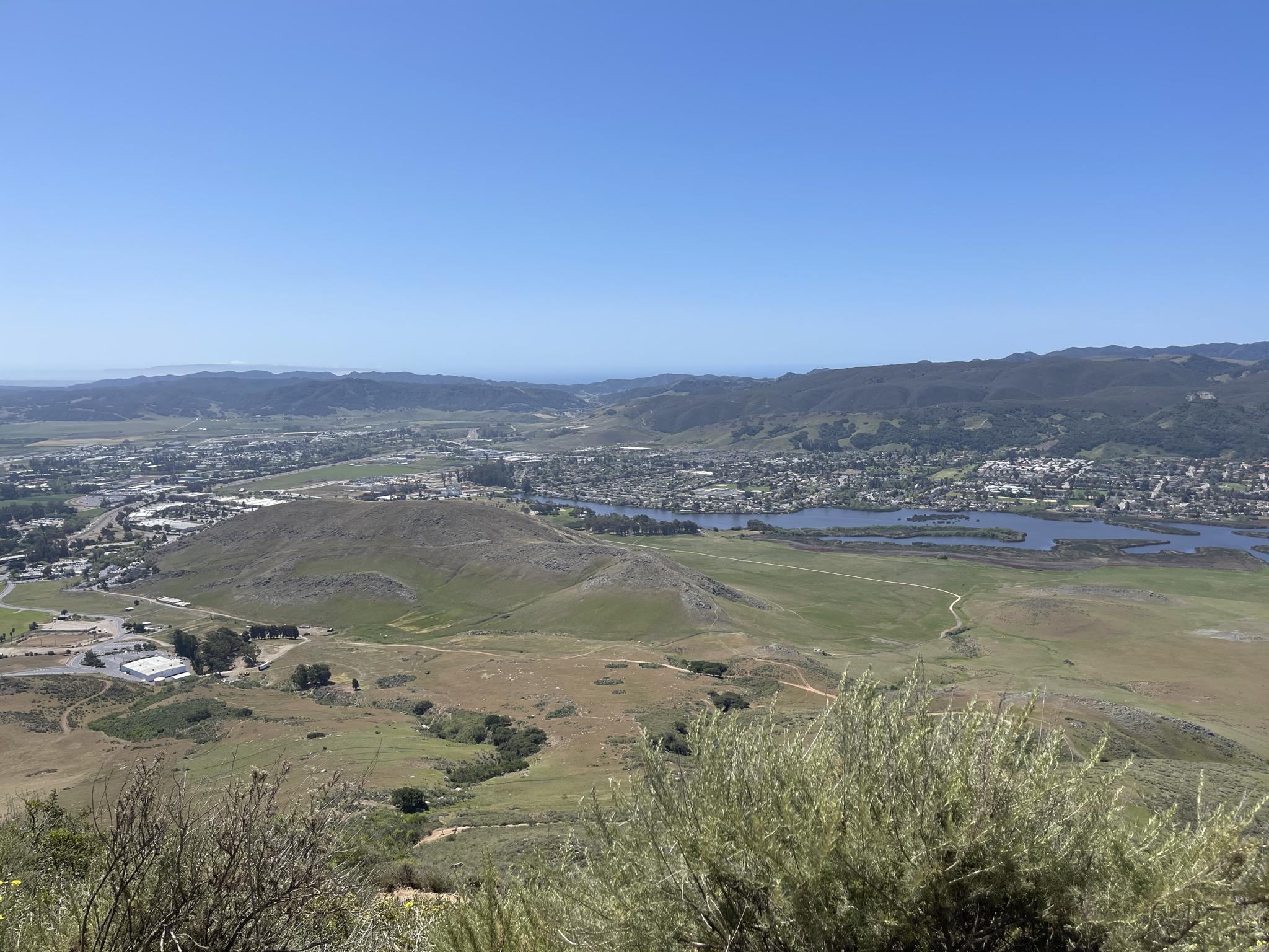 Expansive valley view with a town, water, and green hills under clear sky.