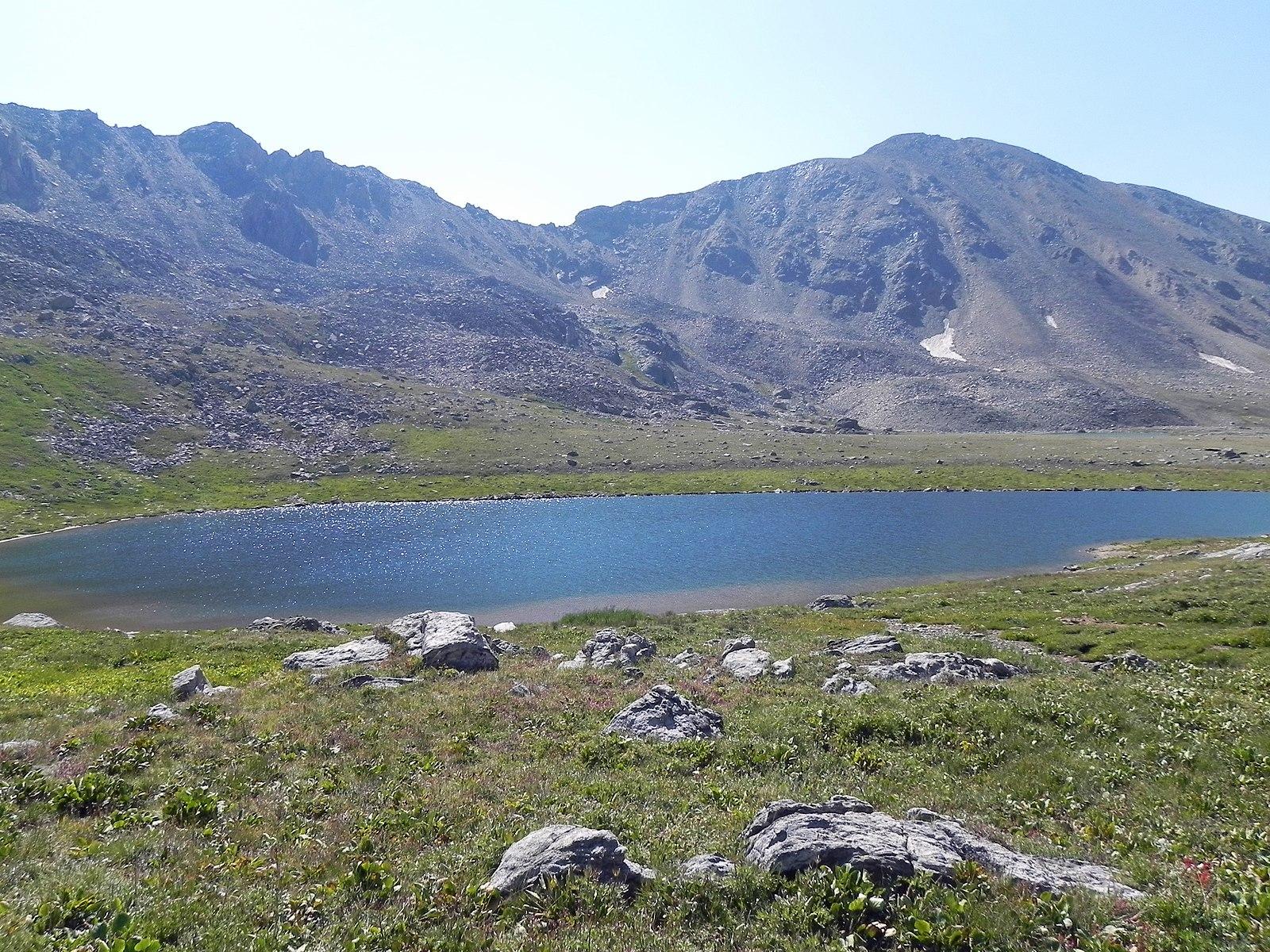 Bright alpine lake surrounded by green grass and rocky mountains under clear skies.