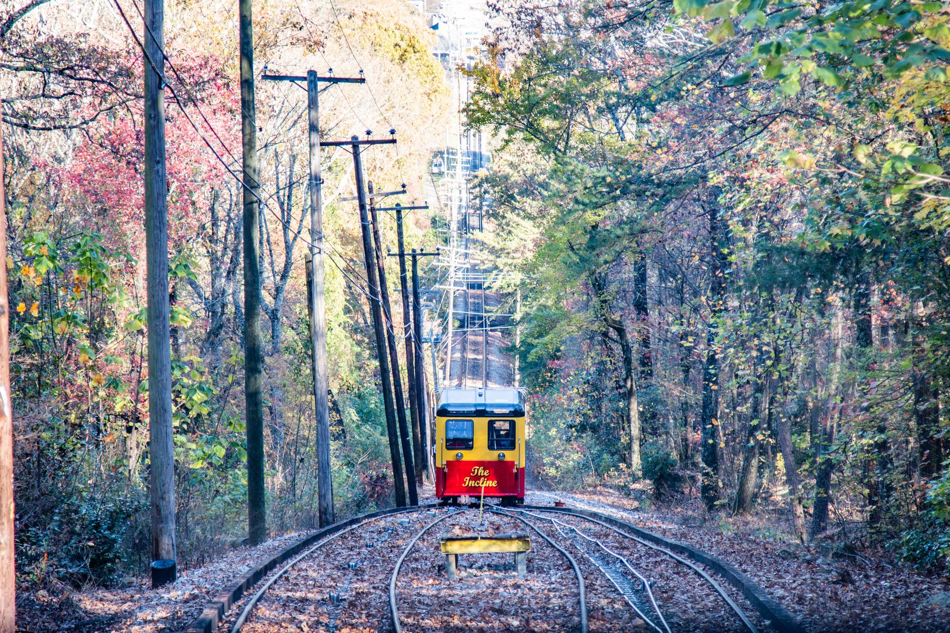 Red and yellow funicular car on tracks ascending a wooded hillside in autumn.