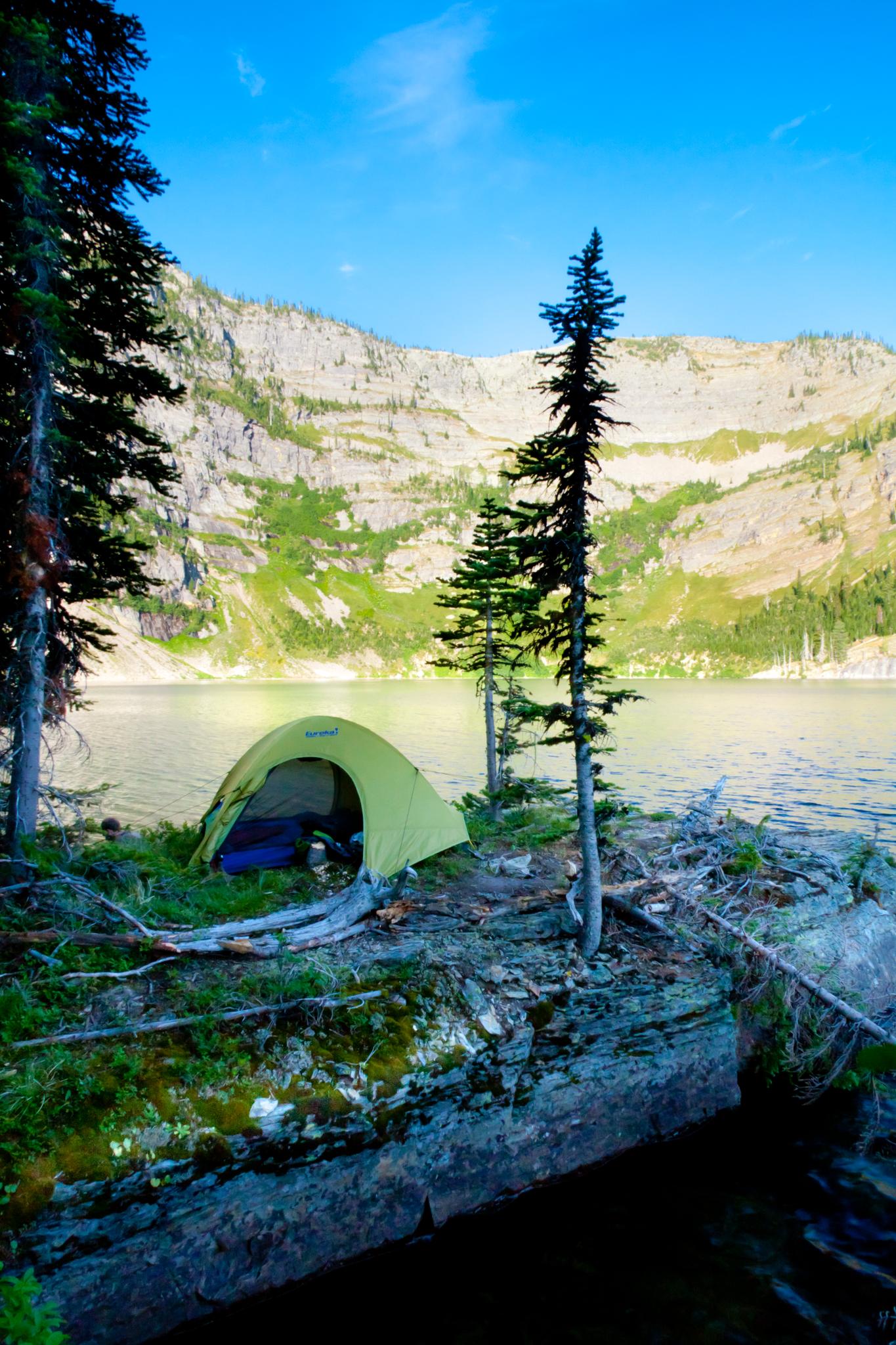 A green tent pitched by a calm lake with mountains under a clear sky.