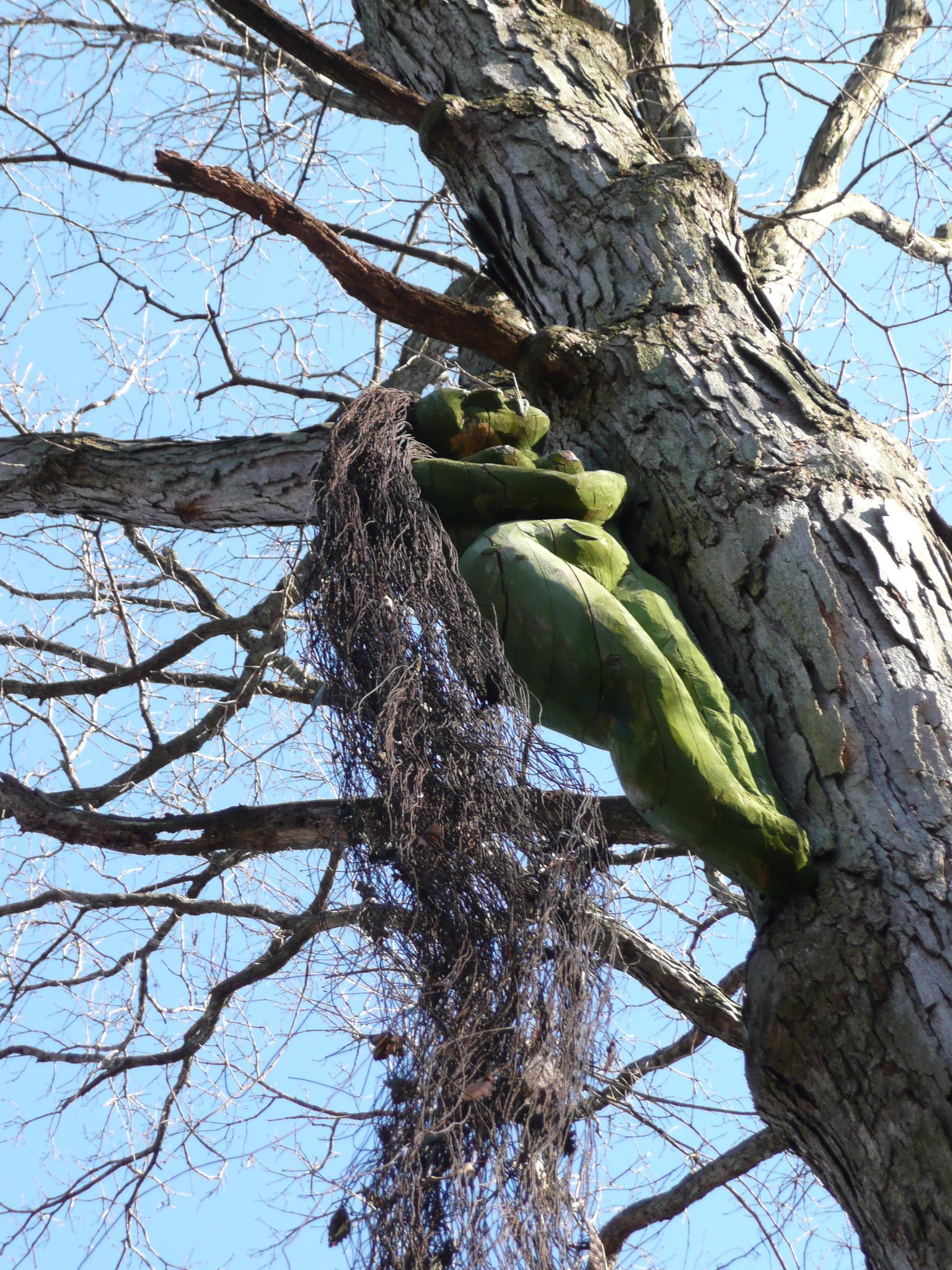 Green mossy figure clinging to a tree trunk with hanging vines under blue sky.