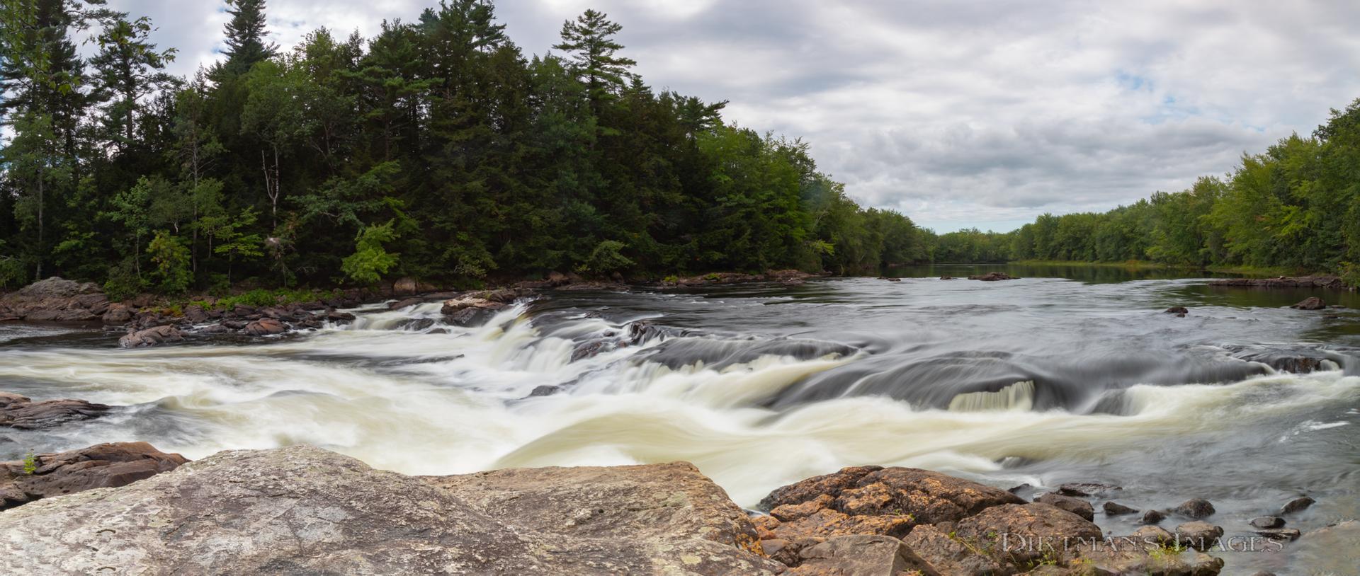 Foaming river rapids cascade through a lush forest under an overcast sky.