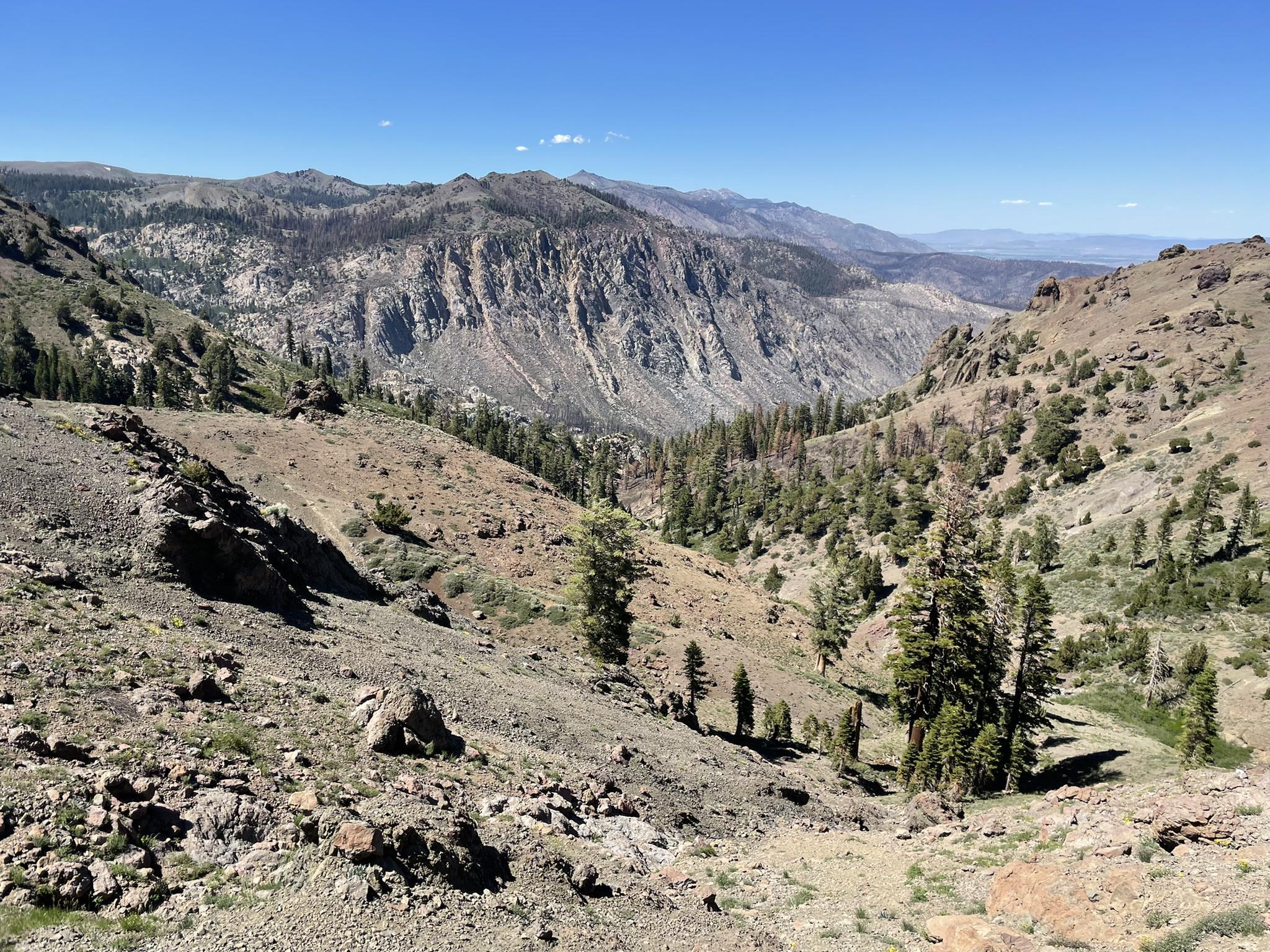 Arid mountain valley with rocky slopes, distant peaks, and sparse trees.
