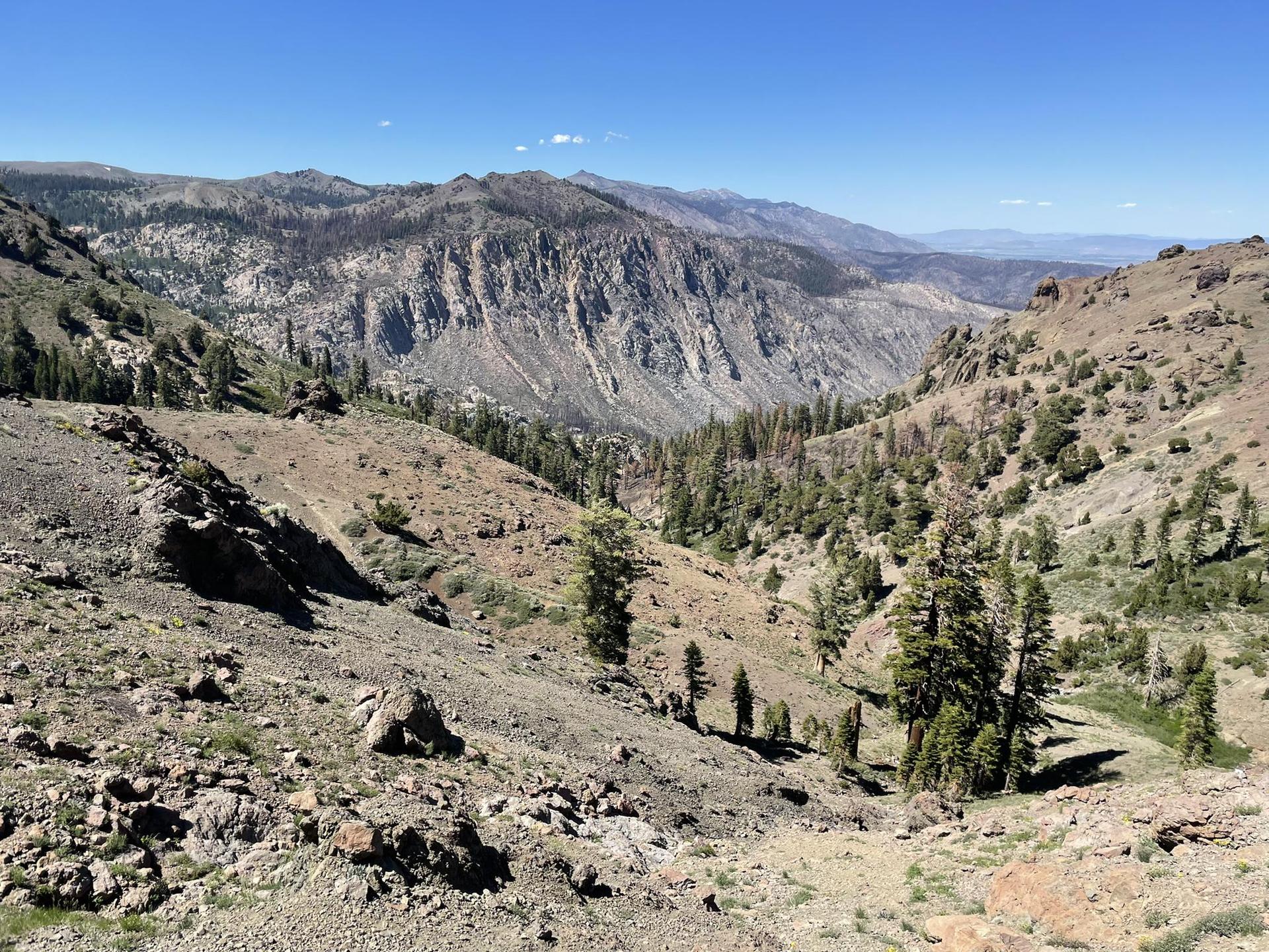 Arid mountain valley with rocky slopes, distant peaks, and sparse trees.
