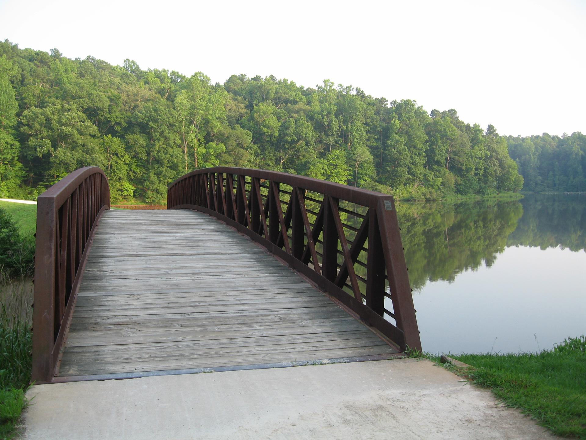 Wooden pedestrian bridge curving over a calm lake with green forest.