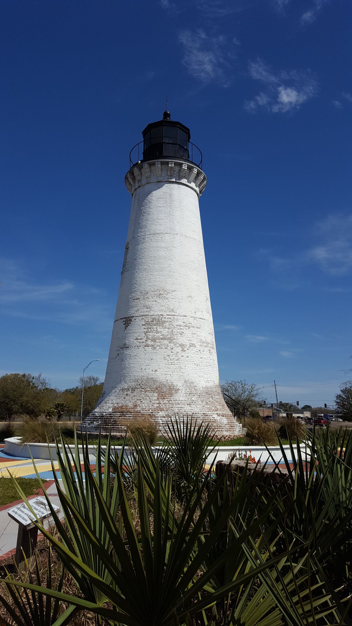 Tall white lighthouse against a bright blue sky, with green foliage in the foreground. - Historic Pascagoula Bike Trail