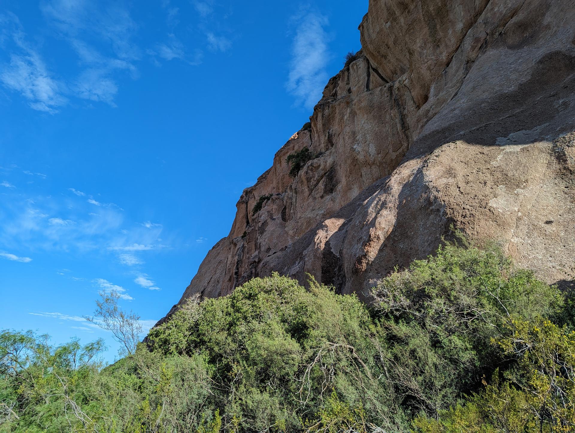 Steep rock face towering over green bushes under a bright blue sky.