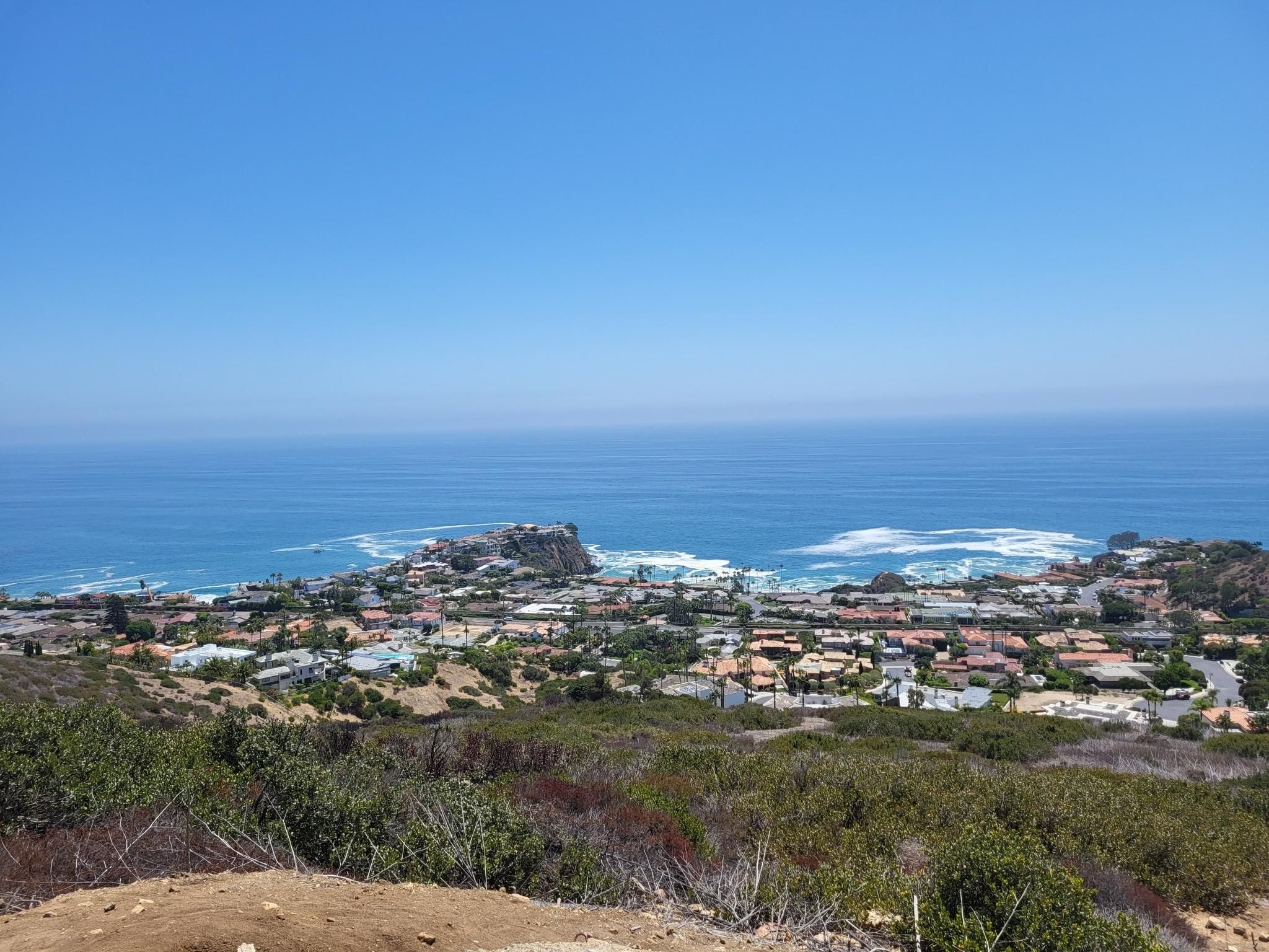 Coastal town nestled by a blue ocean with waves, seen from a dry hillside. - El Moro Canyon Loop Trail