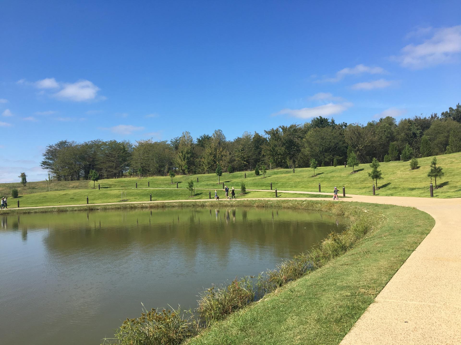 Sunny day, people walk on a path beside a lake and green hills.