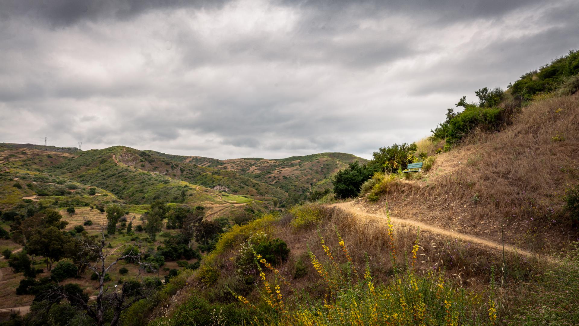 A dirt path winds up a dry hillside, overlooking green valleys under an overcast sky.