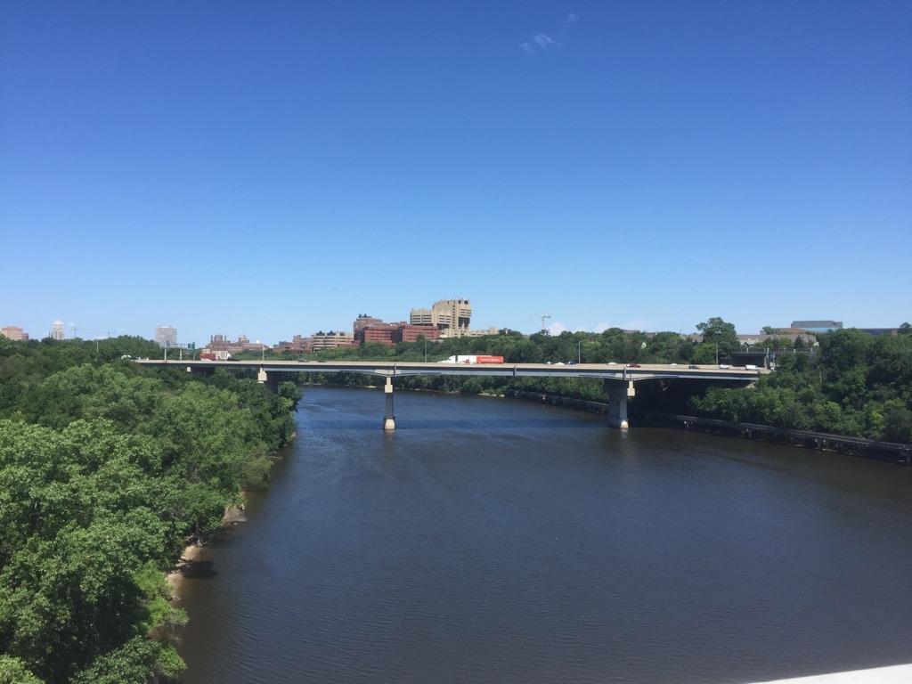 A long bridge spans a wide river, surrounded by green trees under a clear blue sky.
