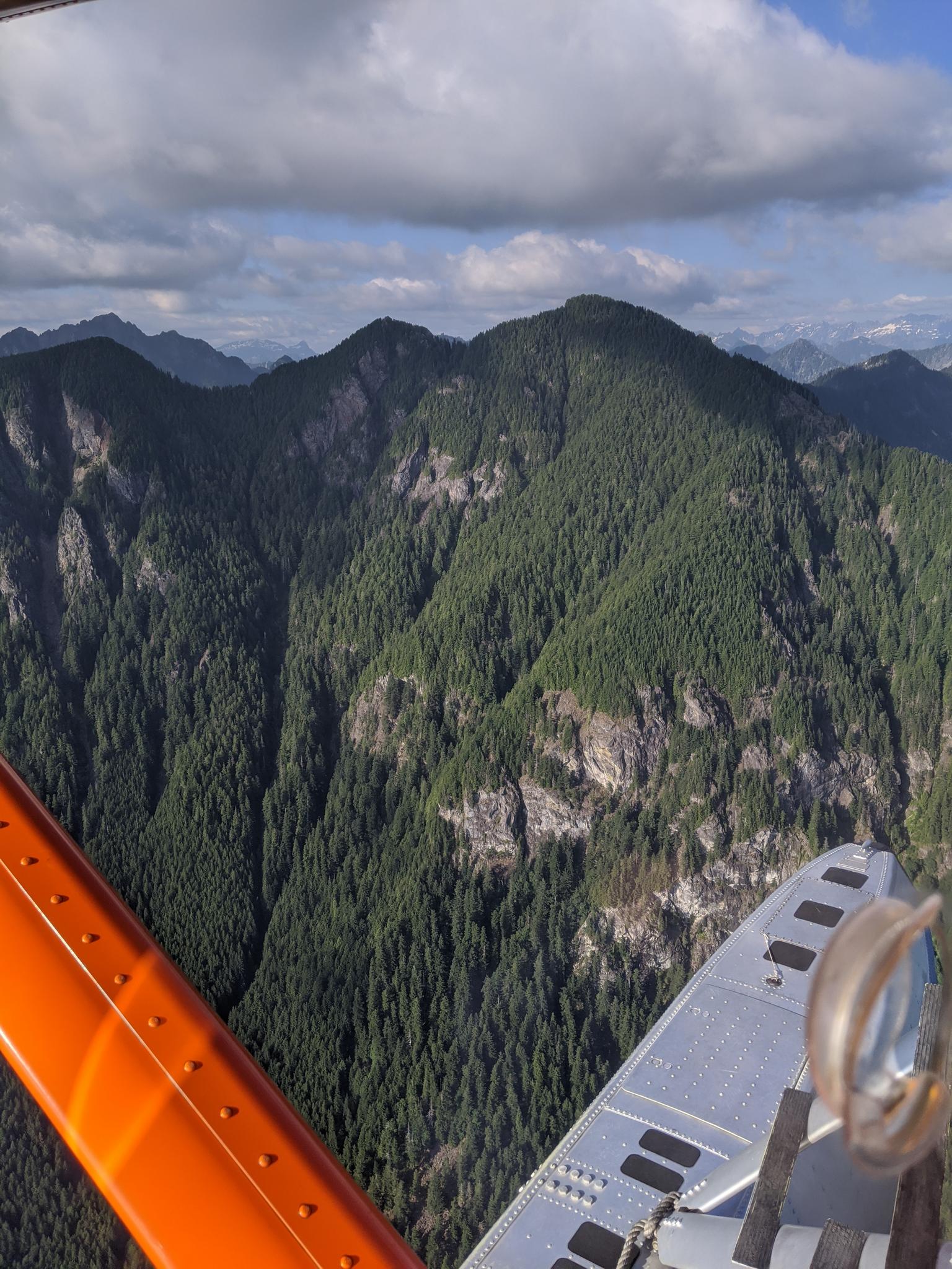 Aerial view of steep, forested mountains under a cloudy sky, airplane wing visible.