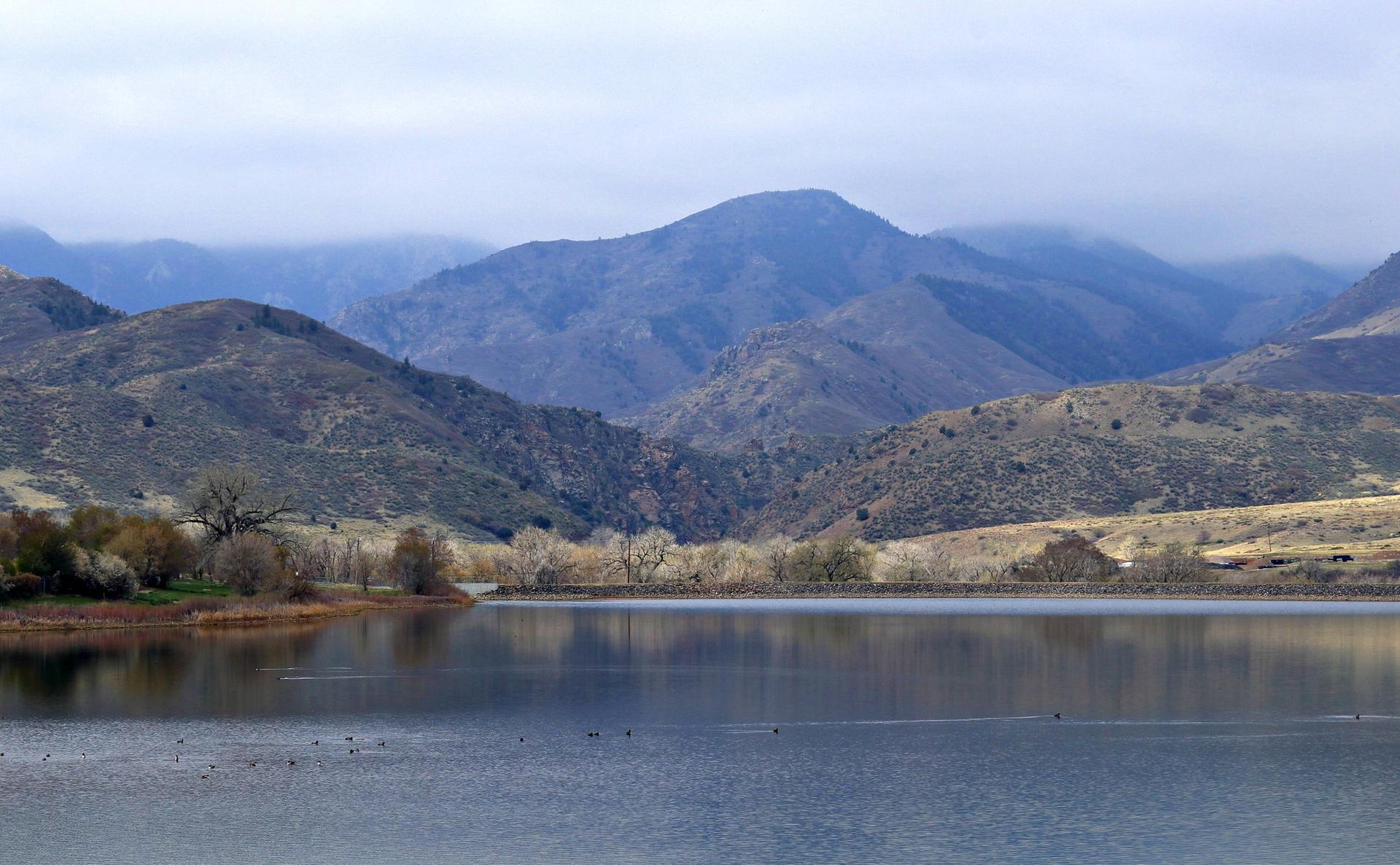 A calm lake reflects cloudy skies, surrounded by bare trees and large mountains.