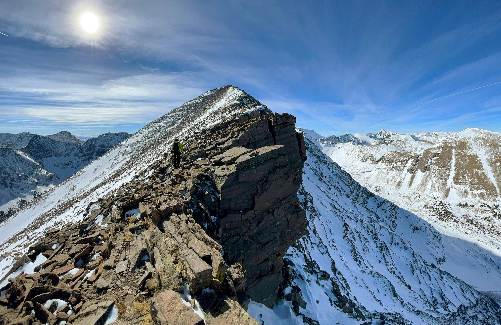 A person hikes a narrow, snowy mountain ridge under a bright sun.