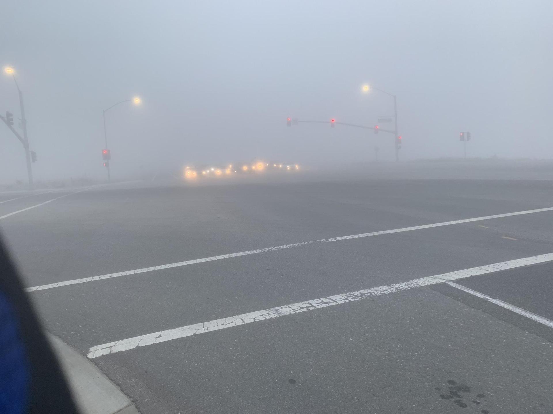 A very foggy intersection with glowing car headlights and traffic lights.