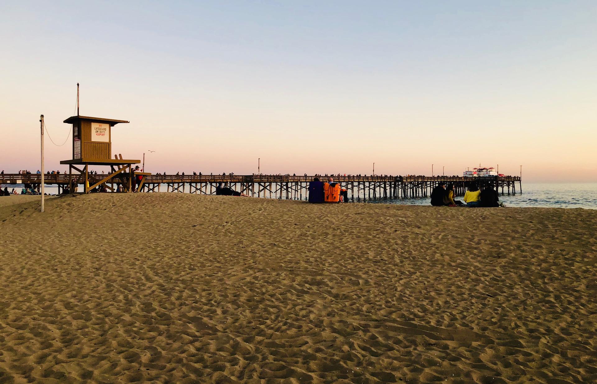 Sandy beach at sunset with a lifeguard tower, pier, and colorful bags.