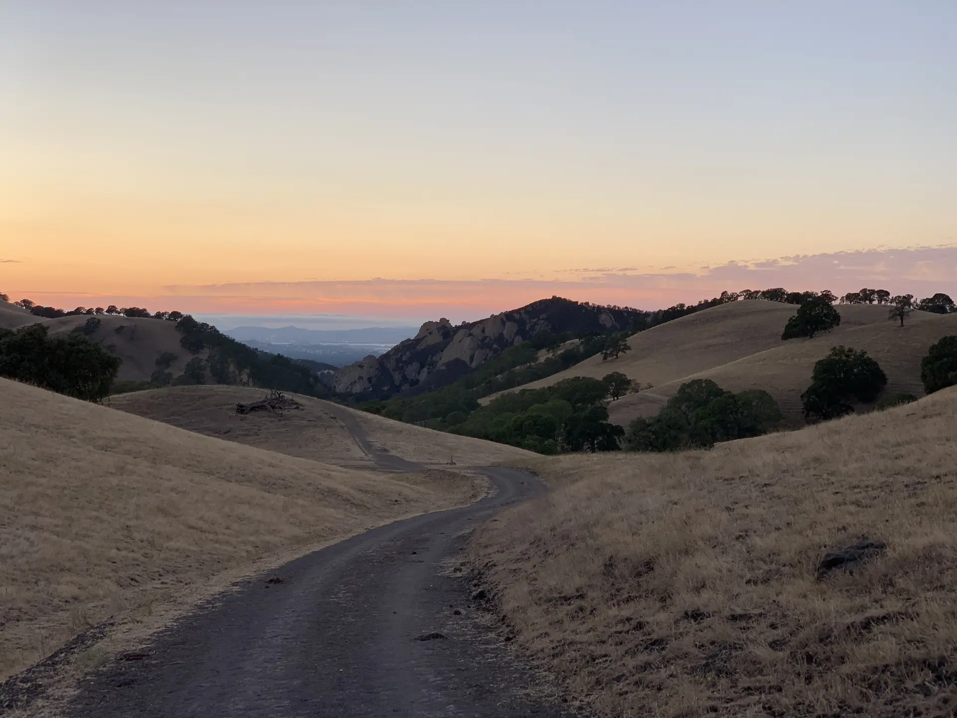 Dirt road through golden hills at sunset with a colorful sky.
