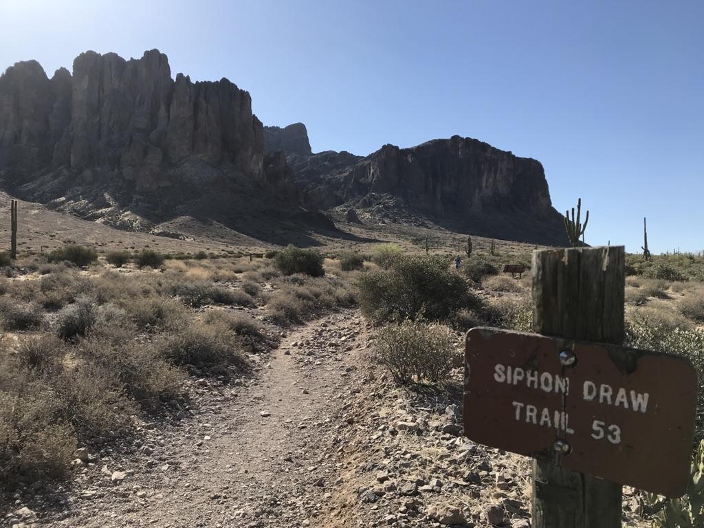 Desert trail sign points towards rugged mountains under a bright sky.