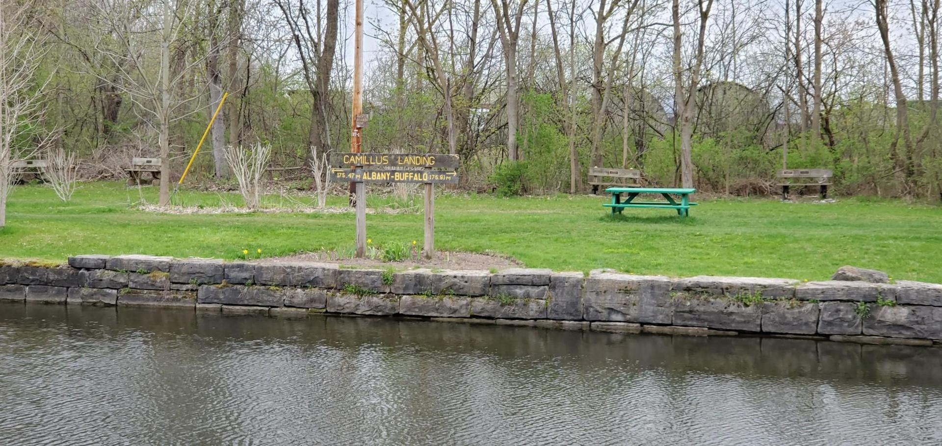 Grassy picnic area with a green table beside a canal, under an overcast sky.