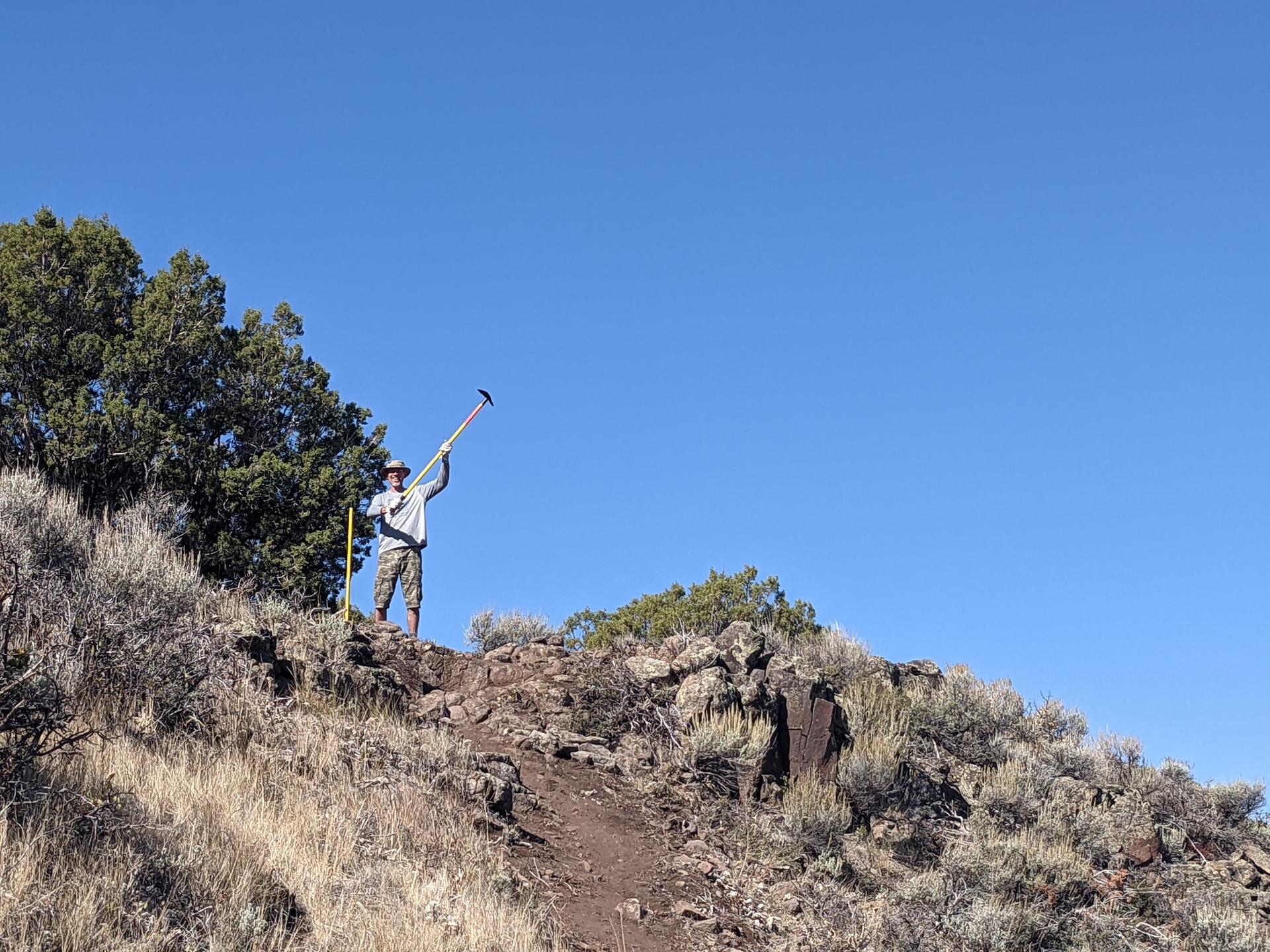 Person holding a long tool on a rocky hillside under a clear blue sky.