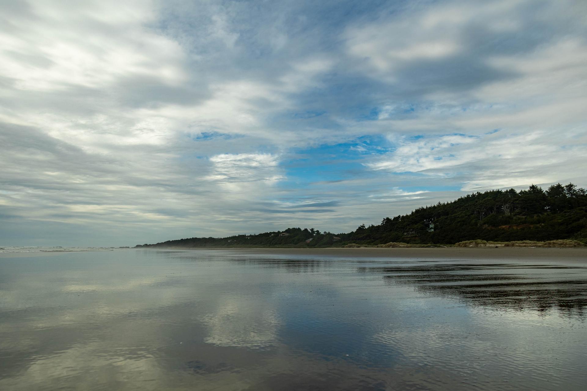 Wide, wet beach reflecting cloudy sky with distant tree-covered hills.