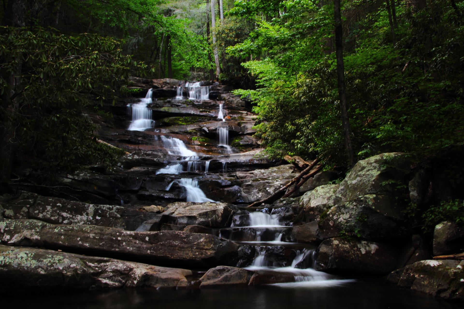 Multi-tiered waterfall cascades over dark rocks into a pool in a lush, shaded forest.