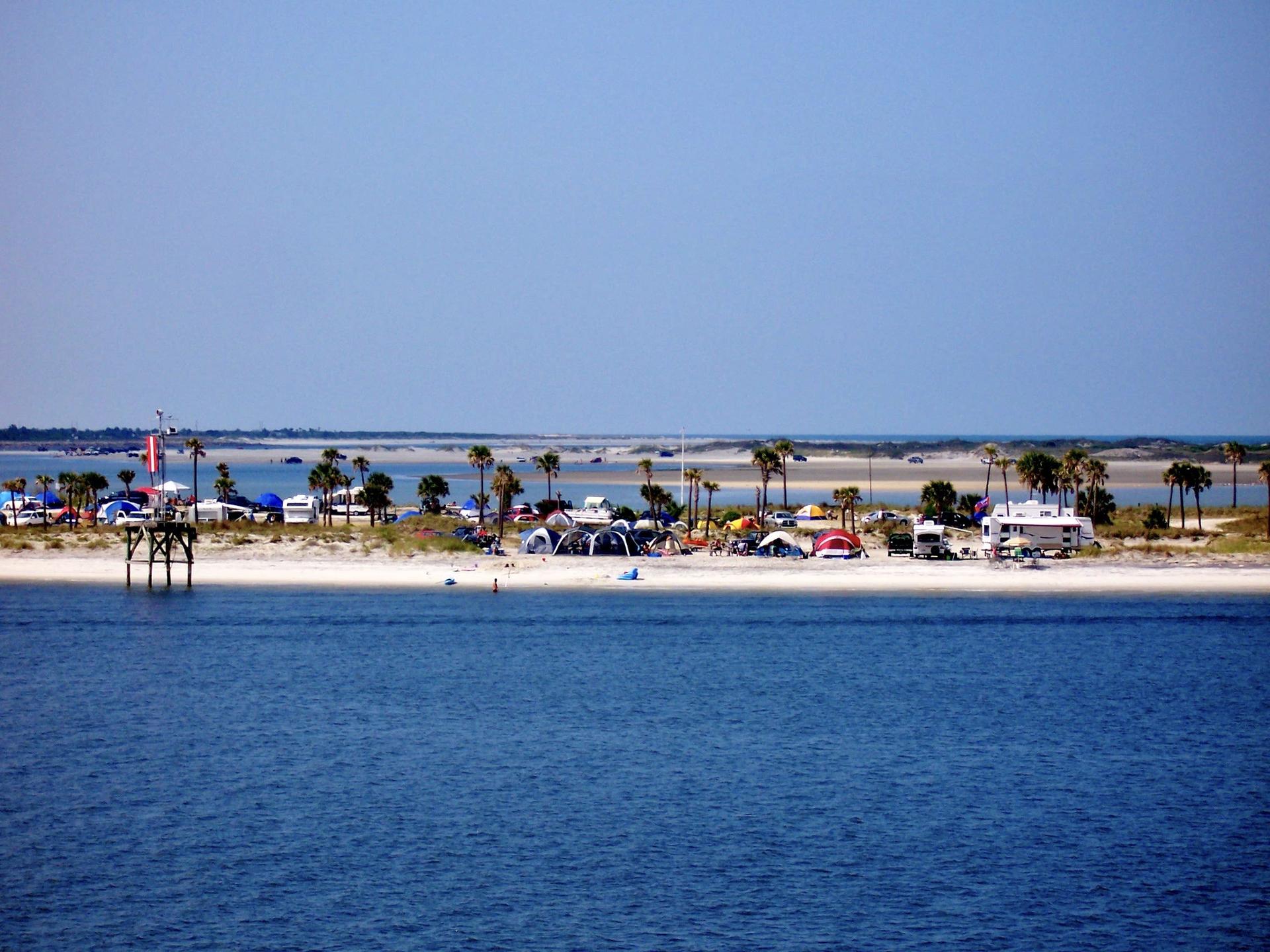 Distant view of a sandy island with many tents, vehicles, and palm trees.