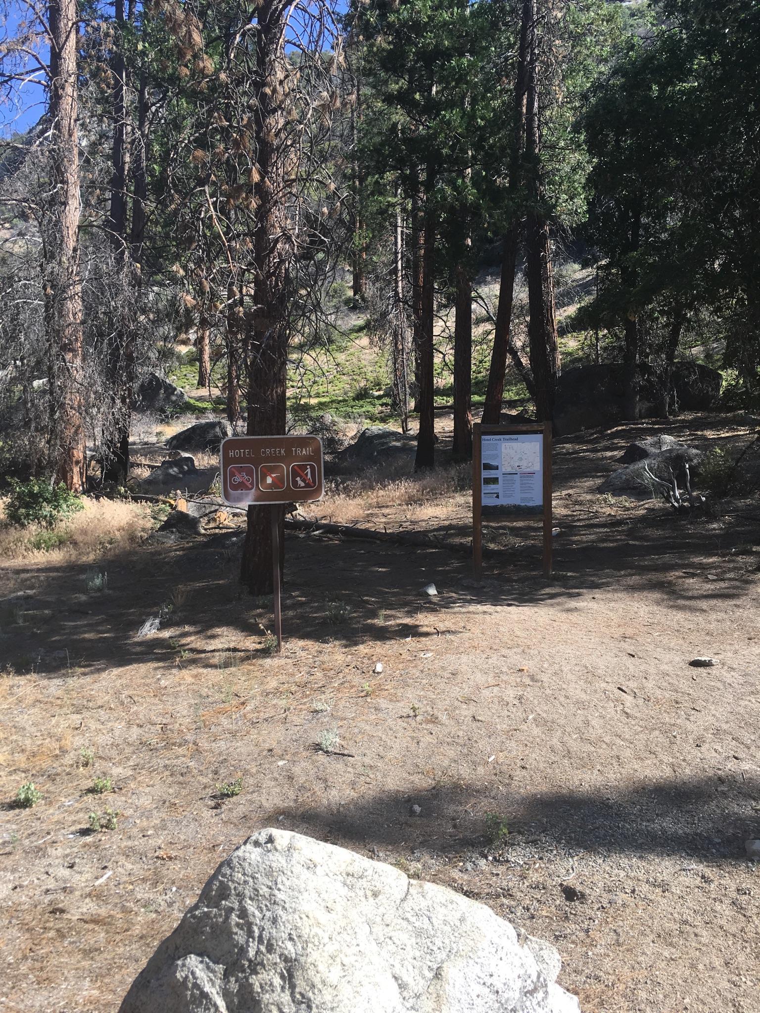 Two trail signs in a dirt clearing surrounded by trees and rocks.
