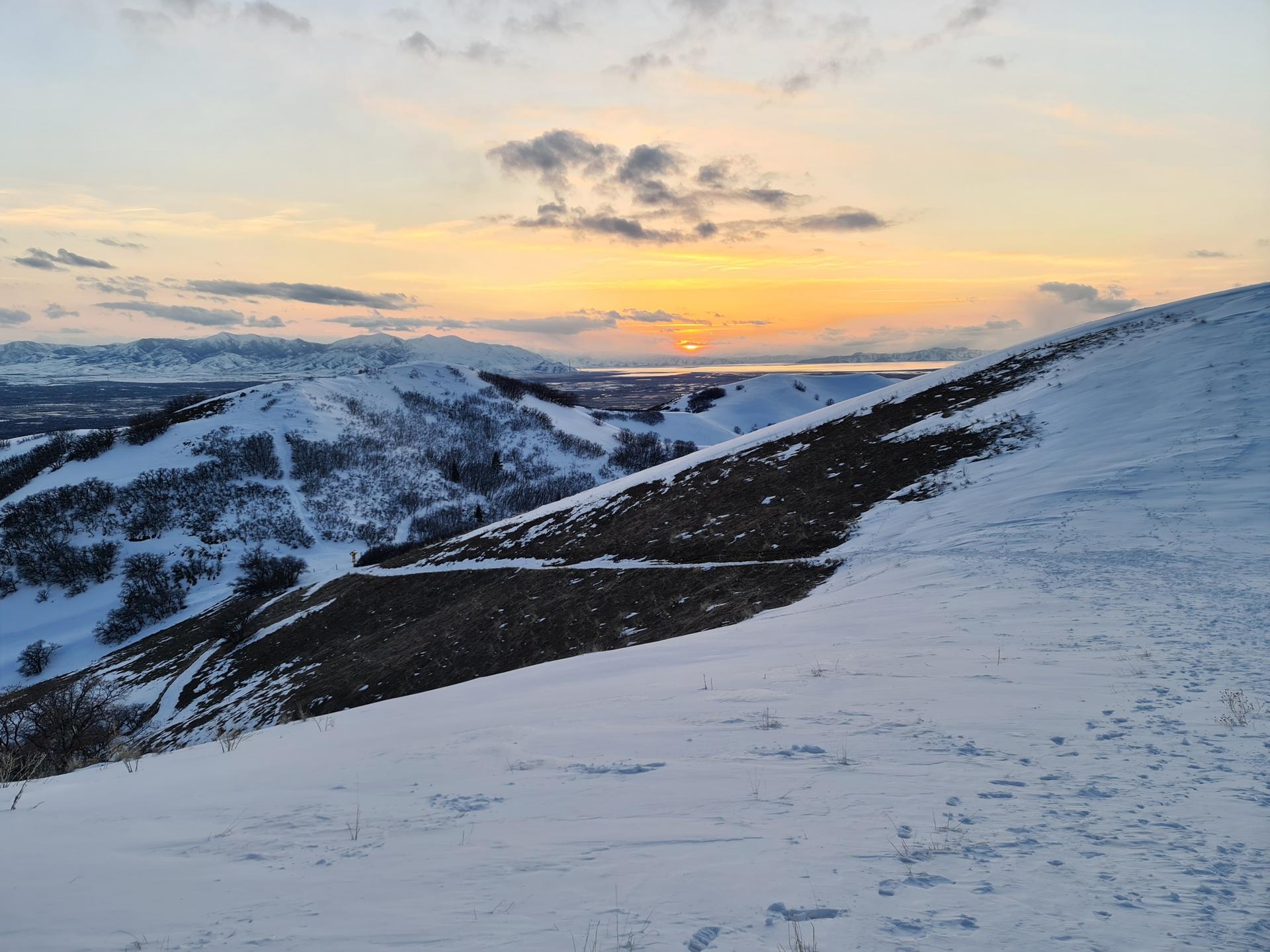 A vibrant sunset over snow-covered hills with dark patches of exposed ground.