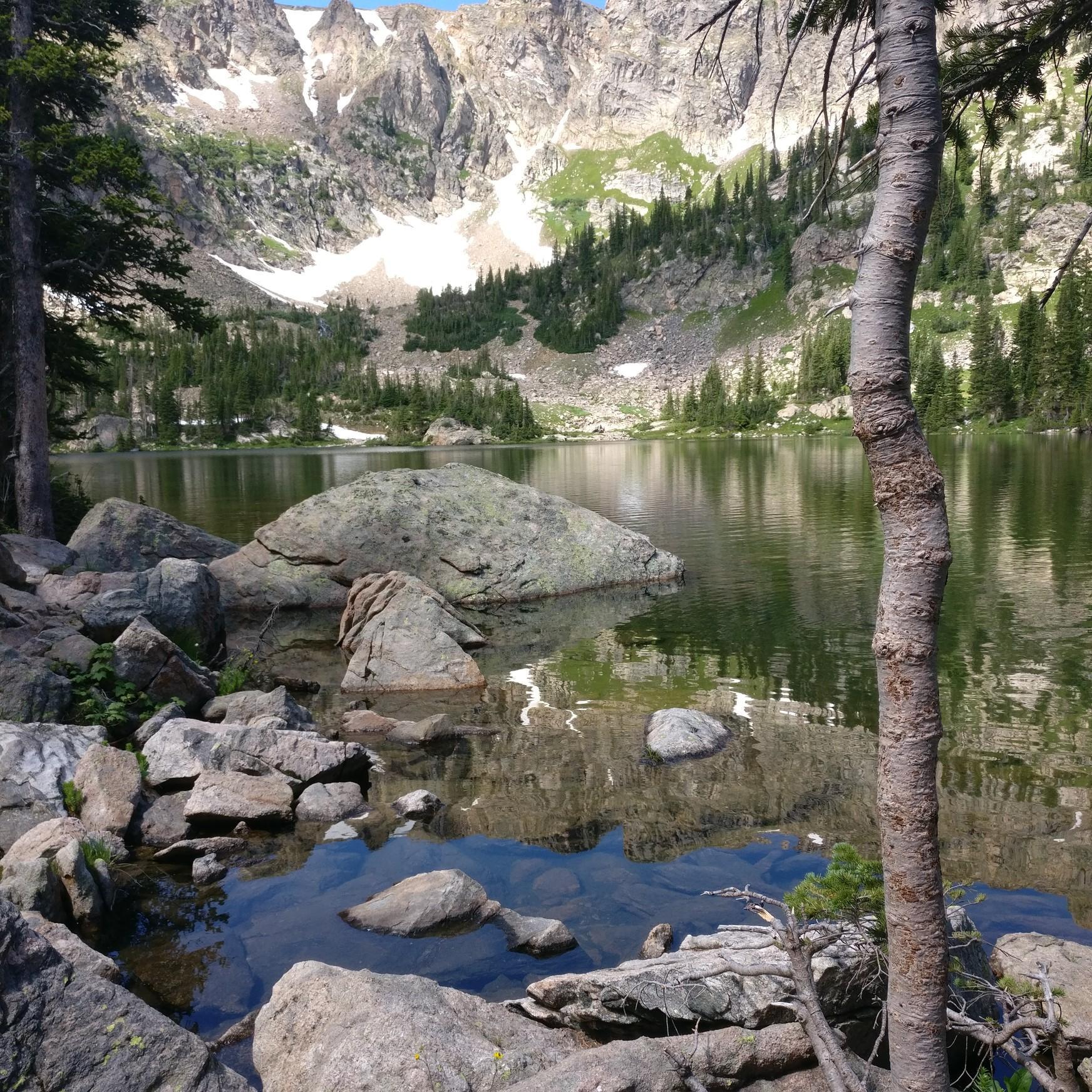 Clear mountain lake reflecting rugged peaks, surrounded by rocks and pine trees.