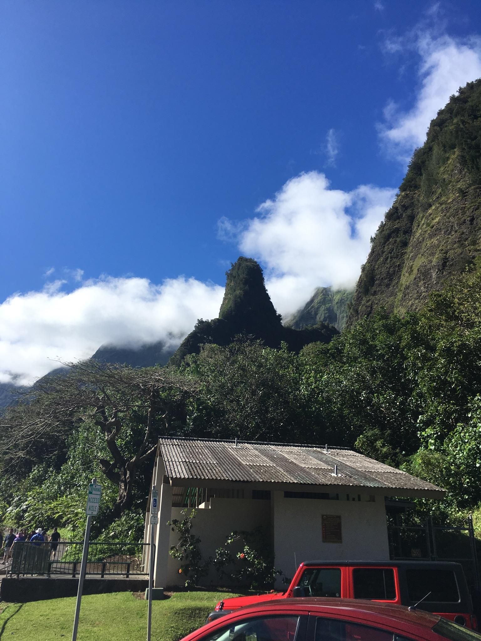 A small building and red car below steep, green mountains on a sunny day. - ʻĪao Needle via Wailuku