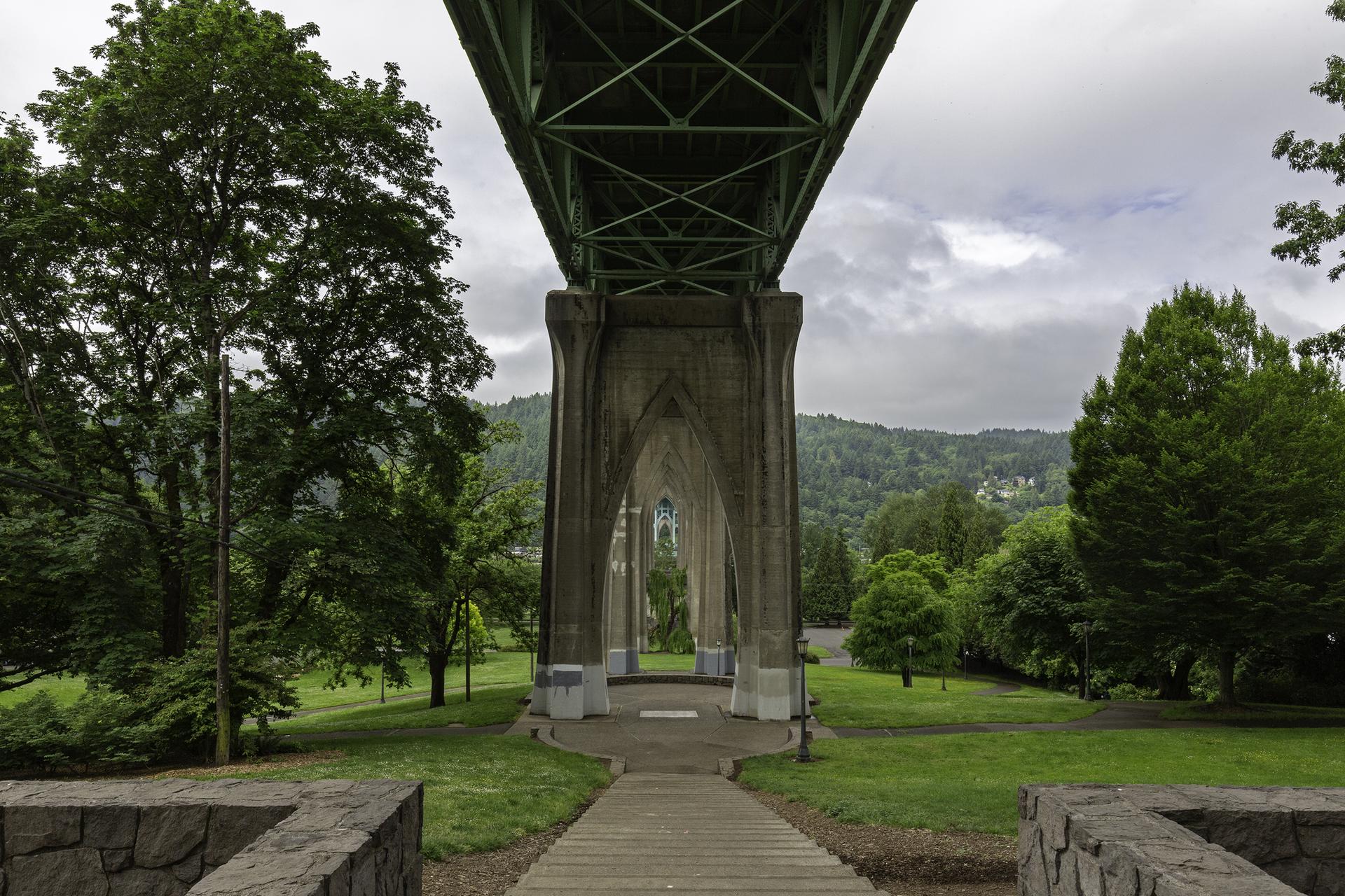 View from under a large bridge with arched supports, green park, and cloudy sky.