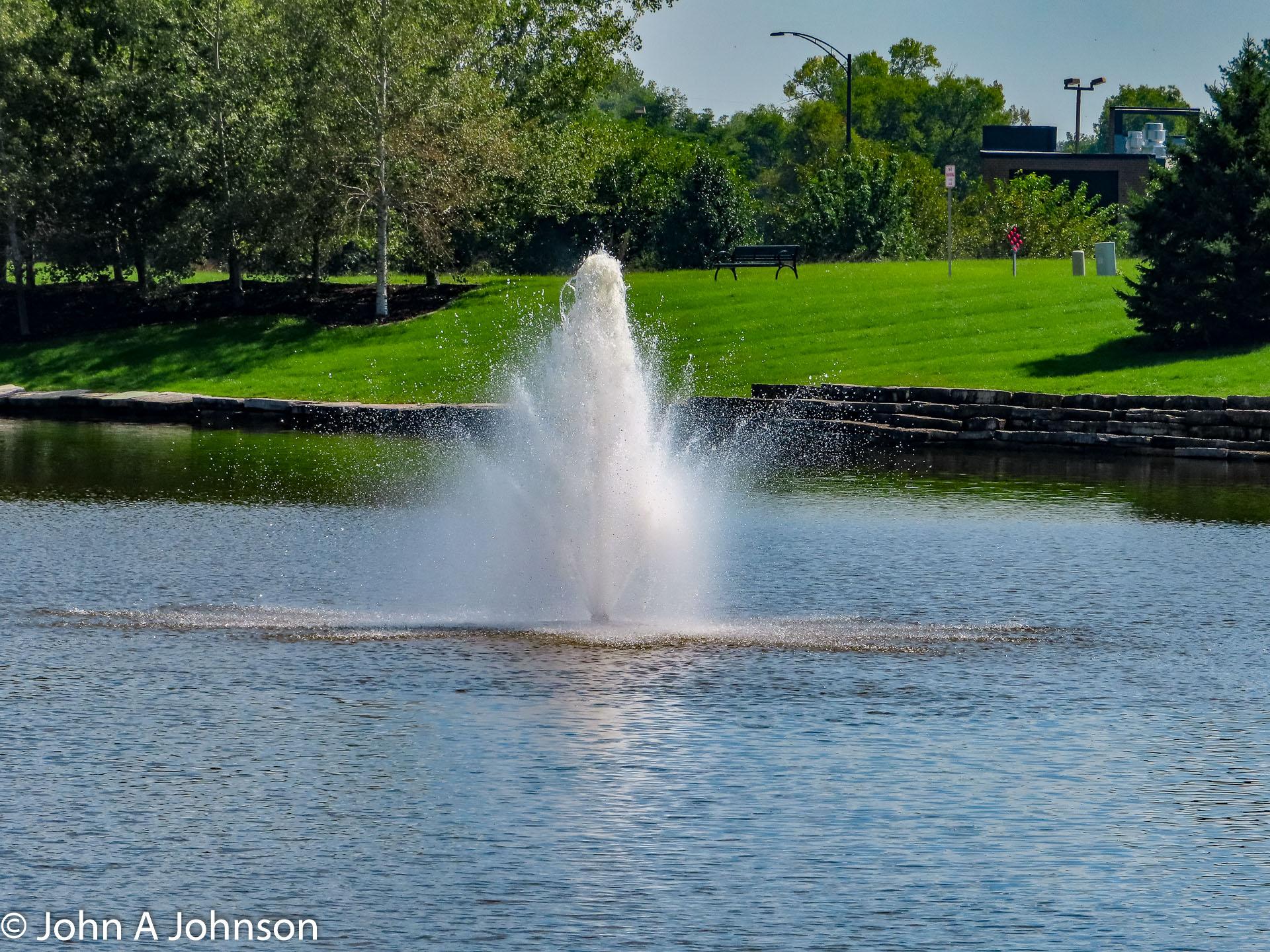 A tall water fountain sprays in a pond with green grass and trees.