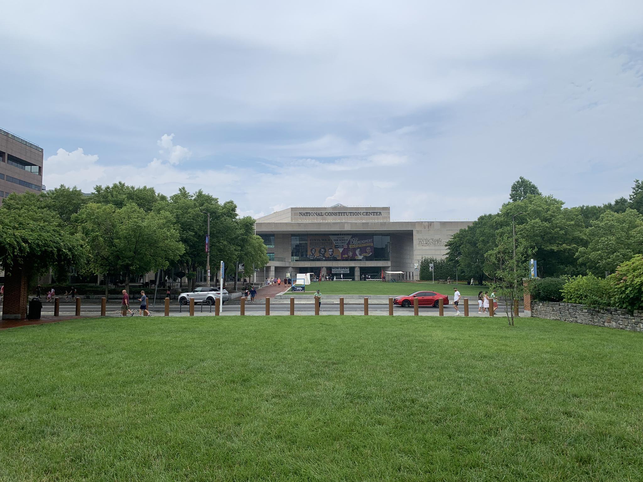 Large modern building behind a green lawn, with cars and people on the street.