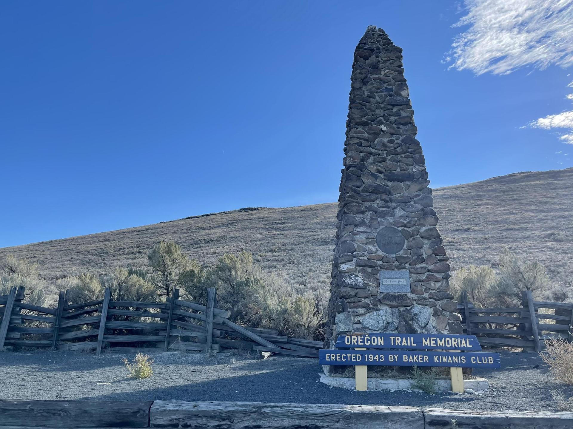 A tall stone monument with a sign, set against a dry hillside and blue sky.