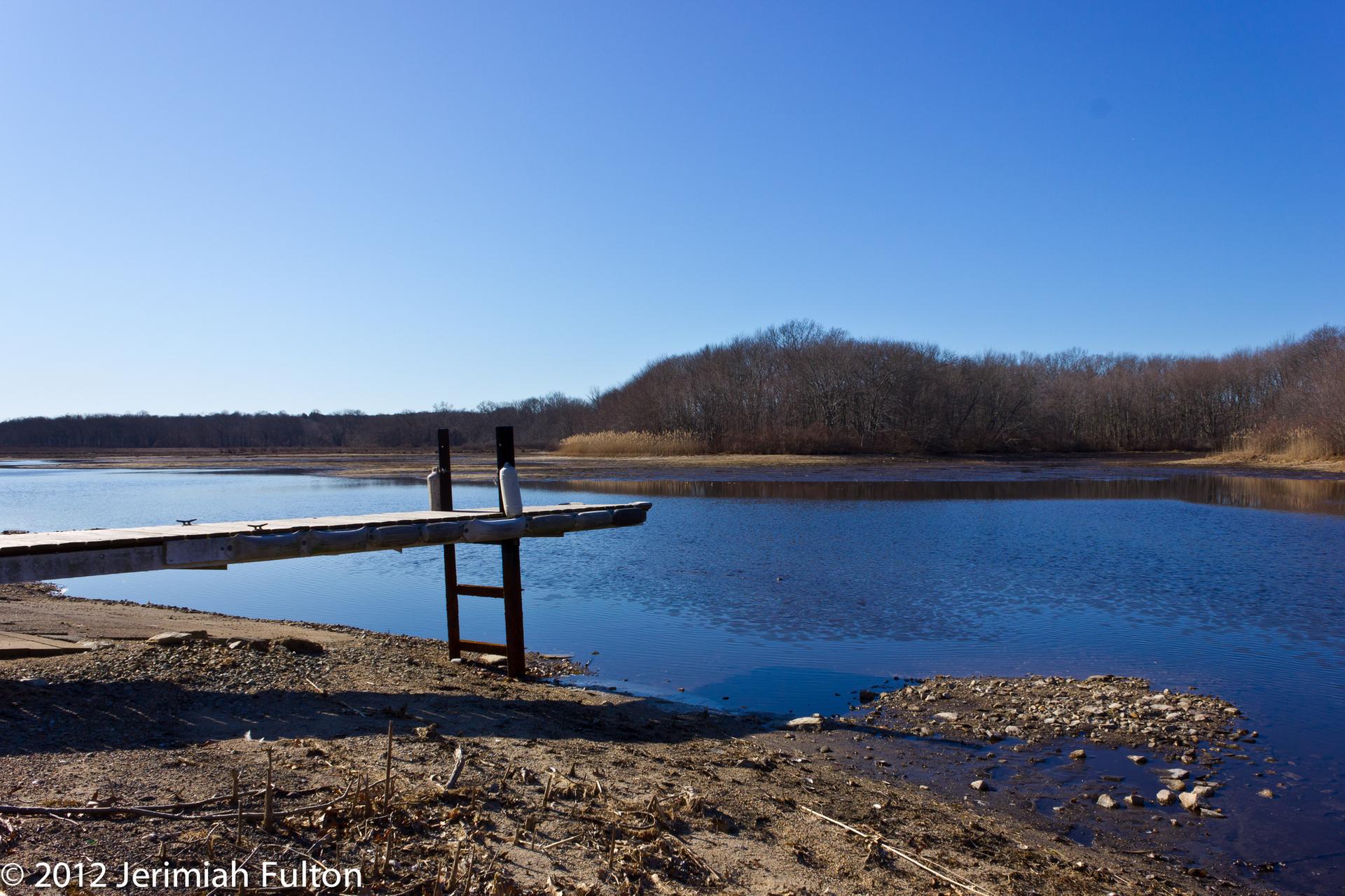 Wooden dock extends into calm water under a clear blue sky. Distant bare trees.