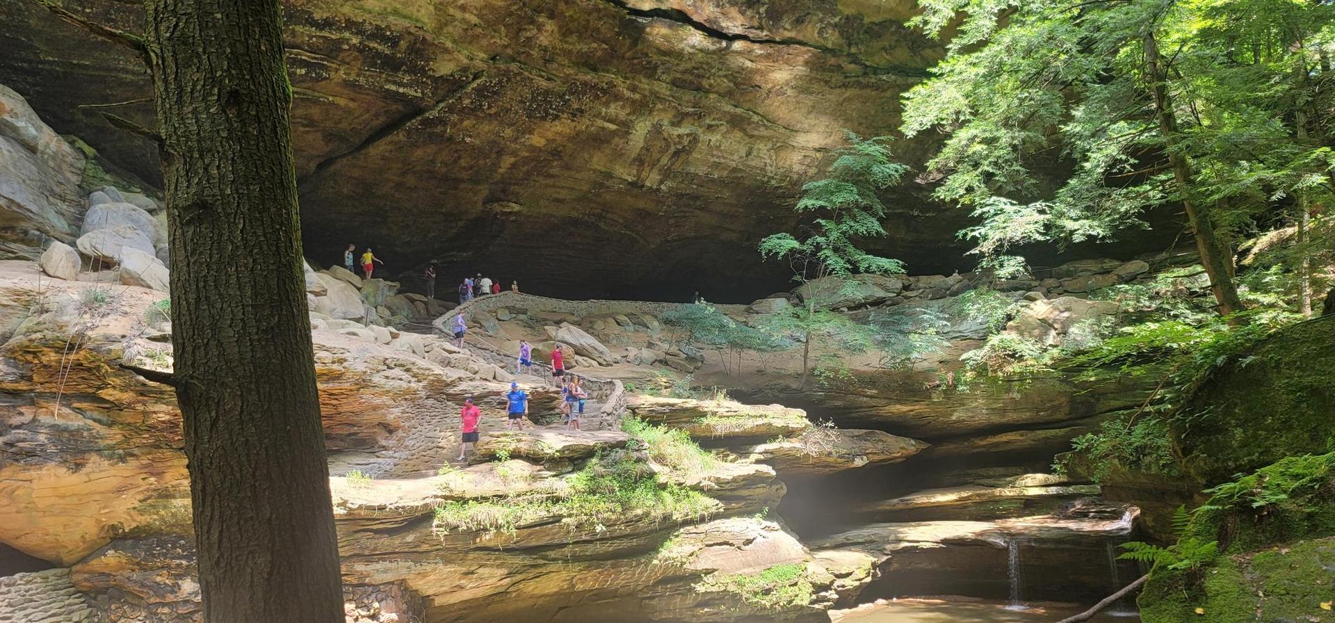 Hikers ascend rocky steps under a massive rock overhang in a green forest.