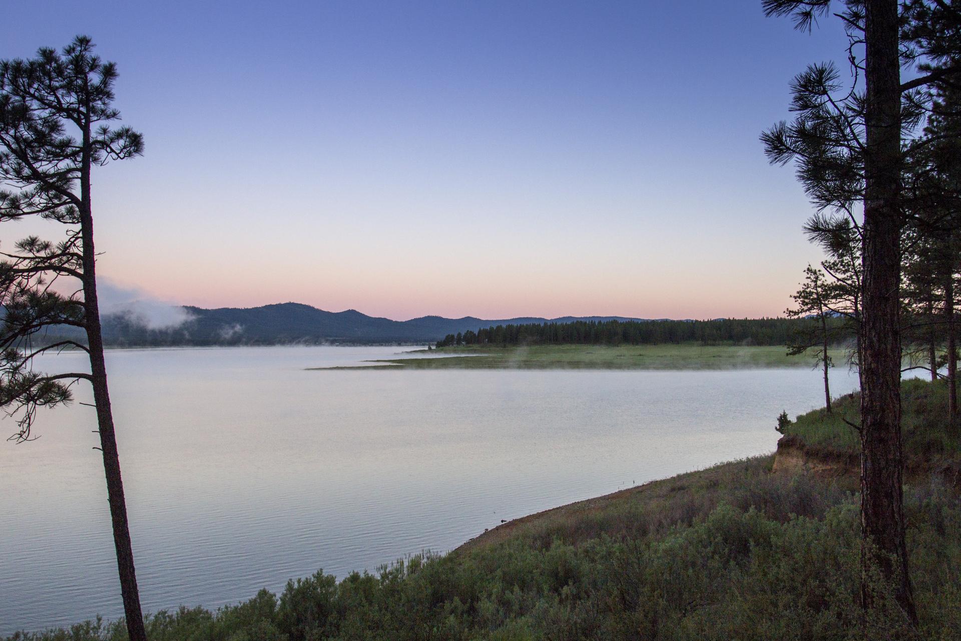 Misty lake at dawn with trees in foreground and mountains in background.